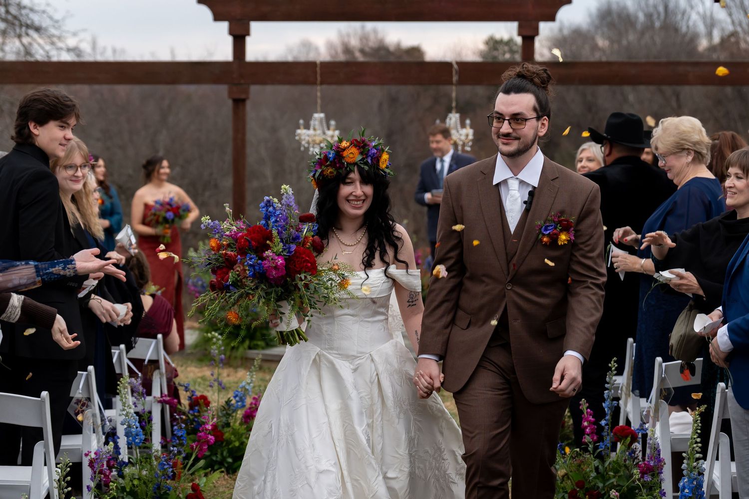 Outdoor wedding ceremony setup with wooden arch and white chairs decorated with colorful flowers.