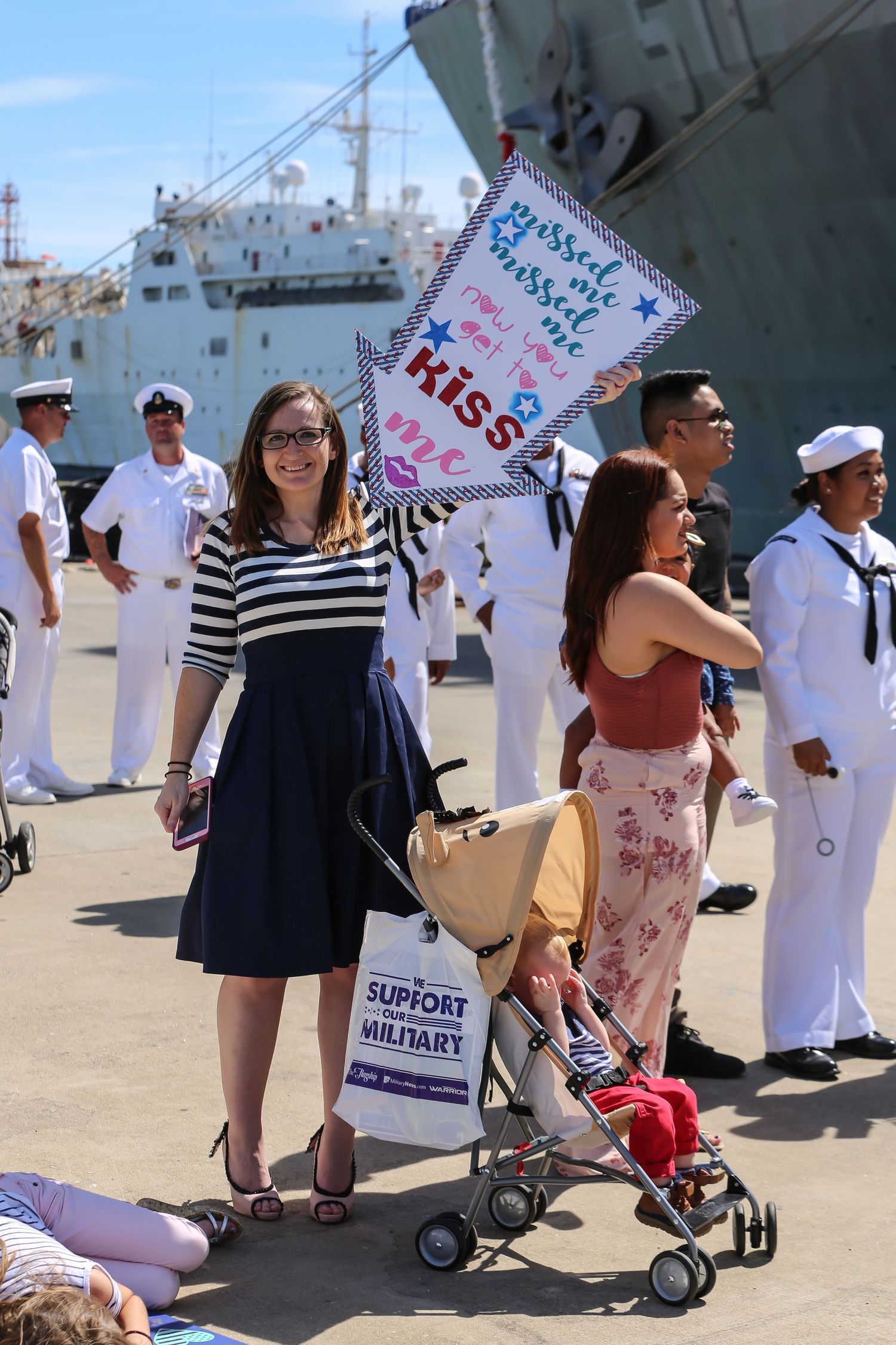 Families with welcome signs gather on dock near naval ship during homecoming celebration.