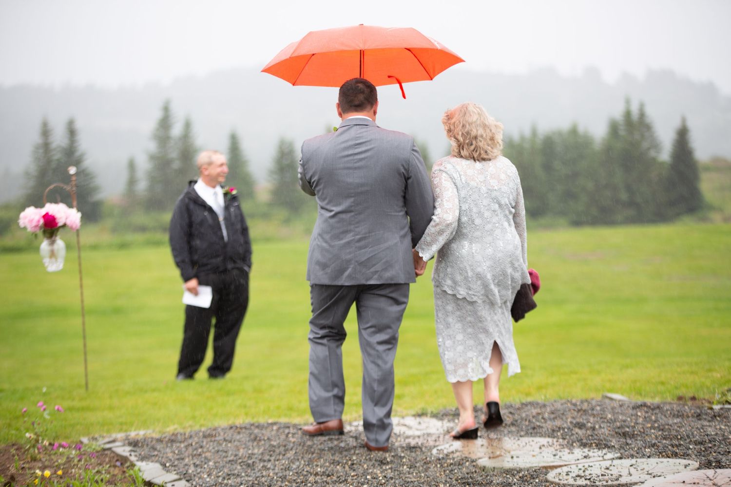 Wedding guests with umbrellas gather on a grey gravel path against a mountain landscape.