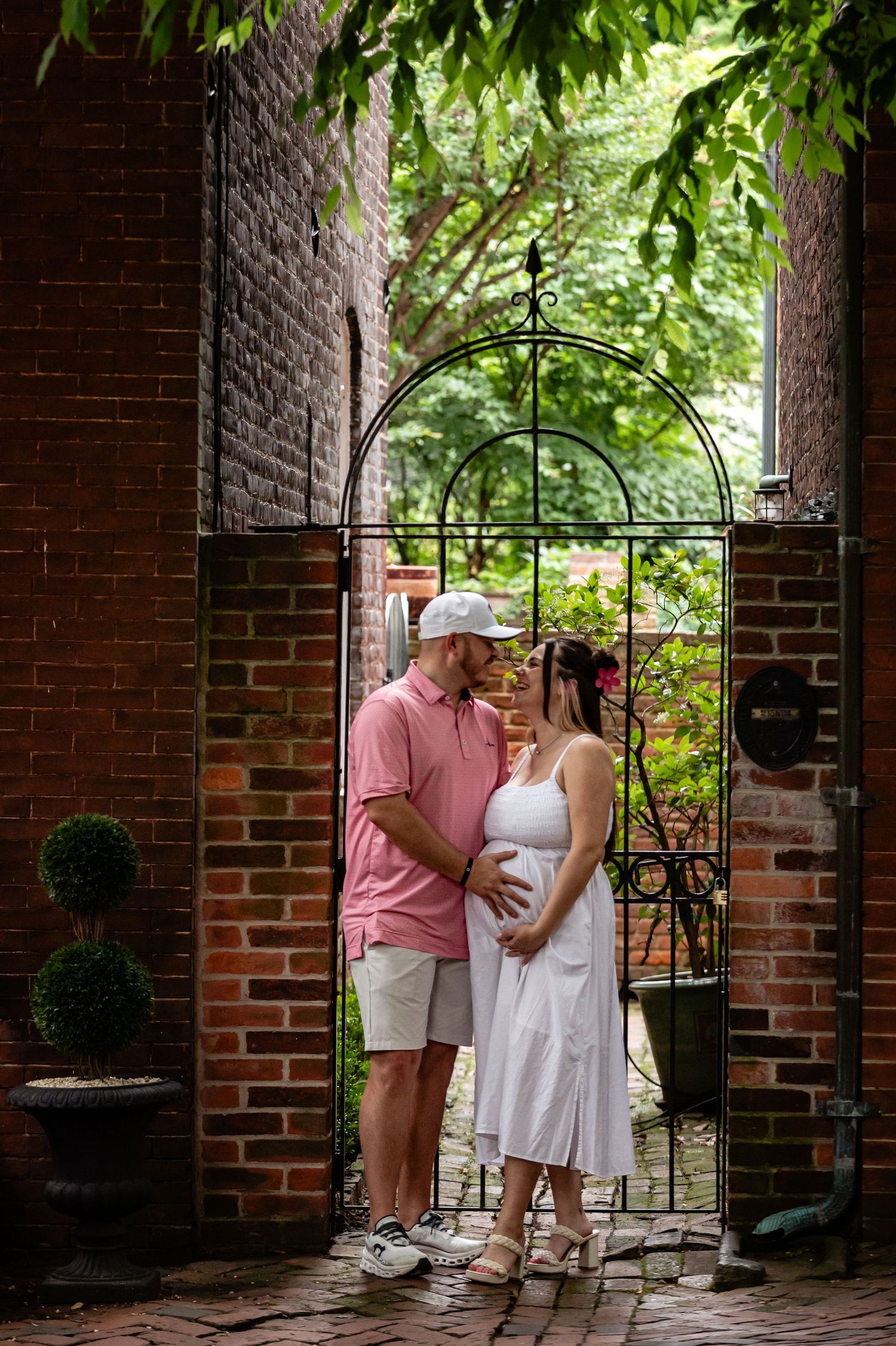 A couple shares a tender moment by a decorative iron gate entrance along a brick wall pathway.