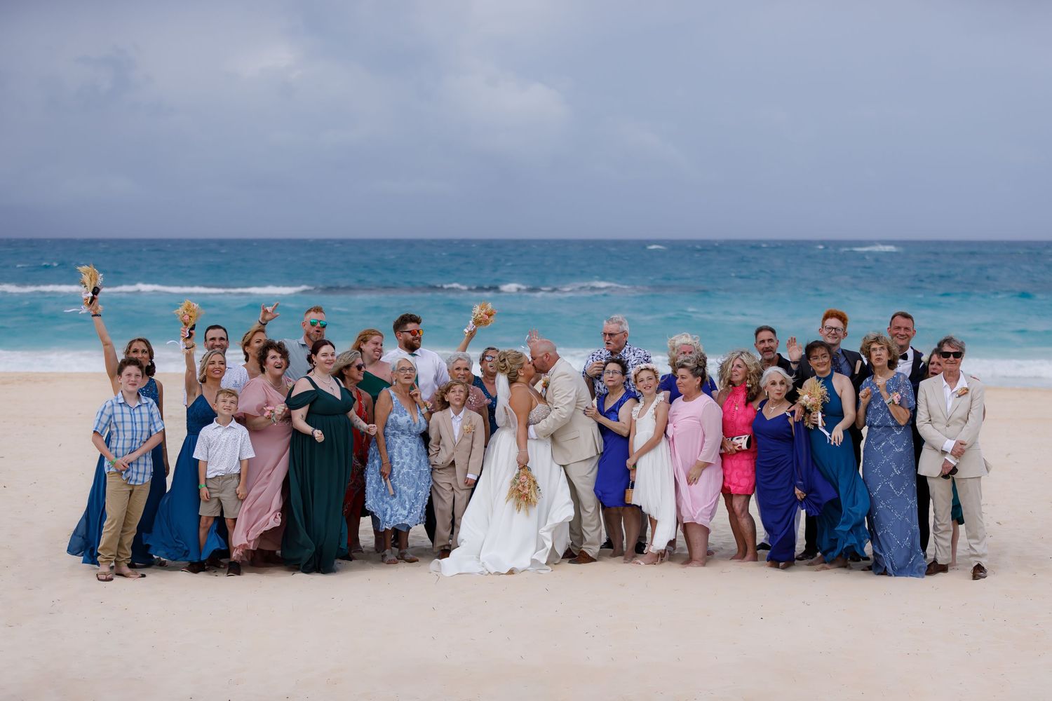 Large wedding party poses together on a tropical beach with turquoise ocean in the background.