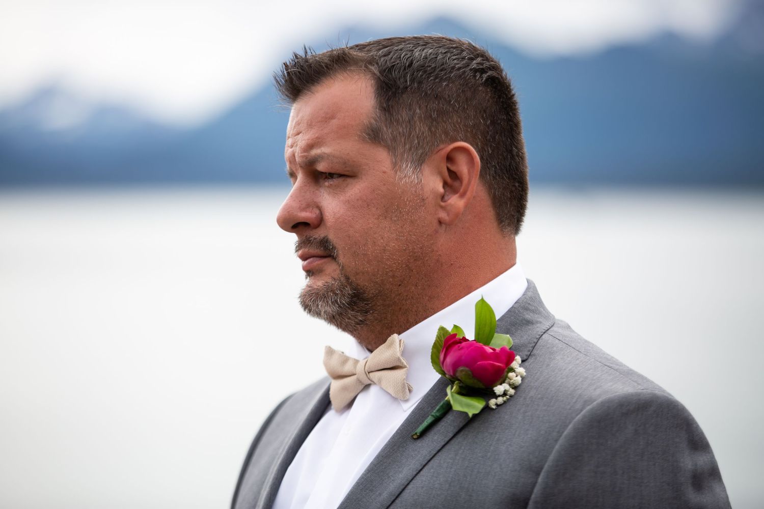 Groomsmen in gray suits and bow ties pose together outdoors with pink flower boutonnieres on a cloudy day.