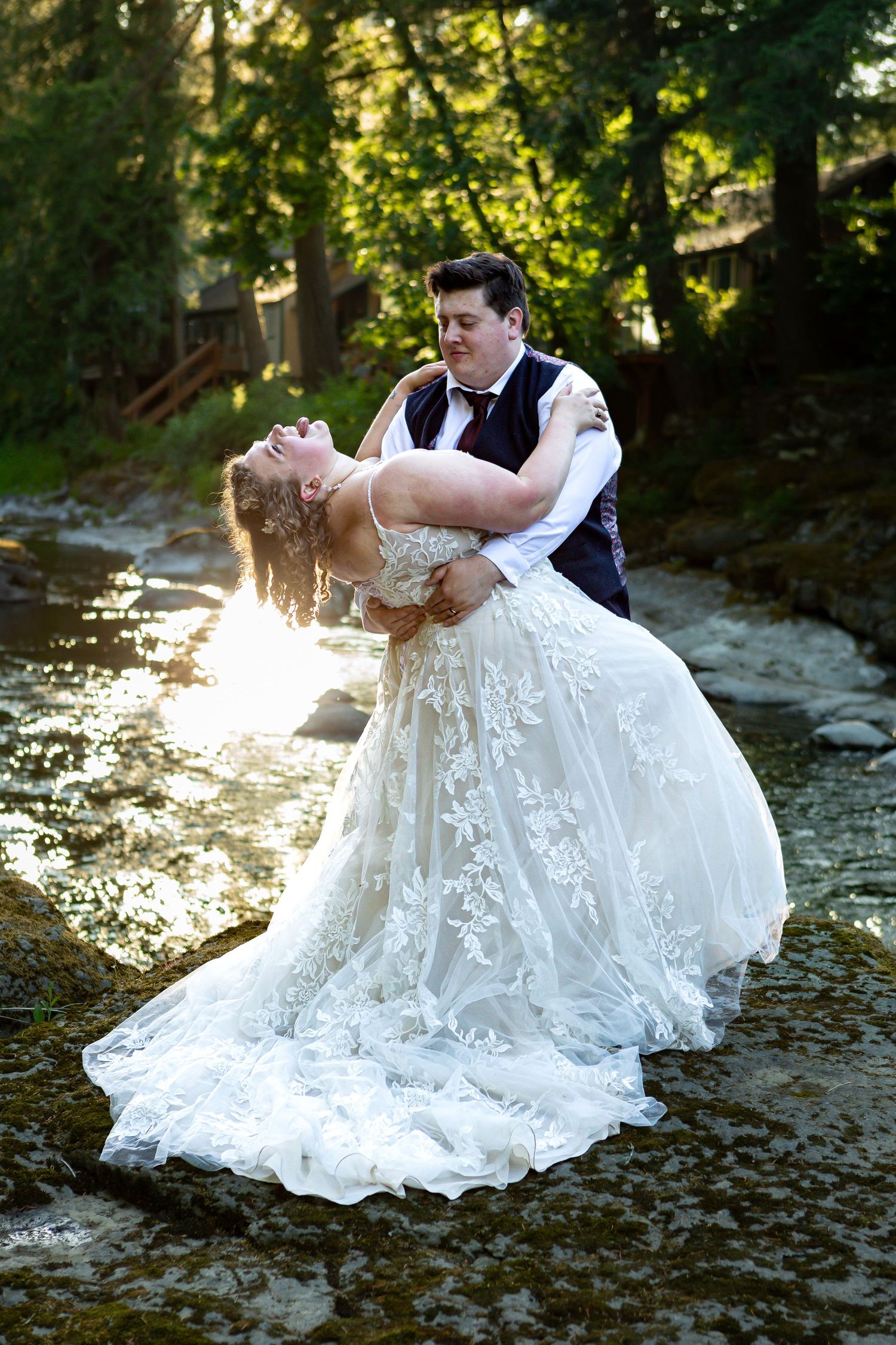 Romantic wedding couple poses dramatically on rocky riverbank at golden hour.