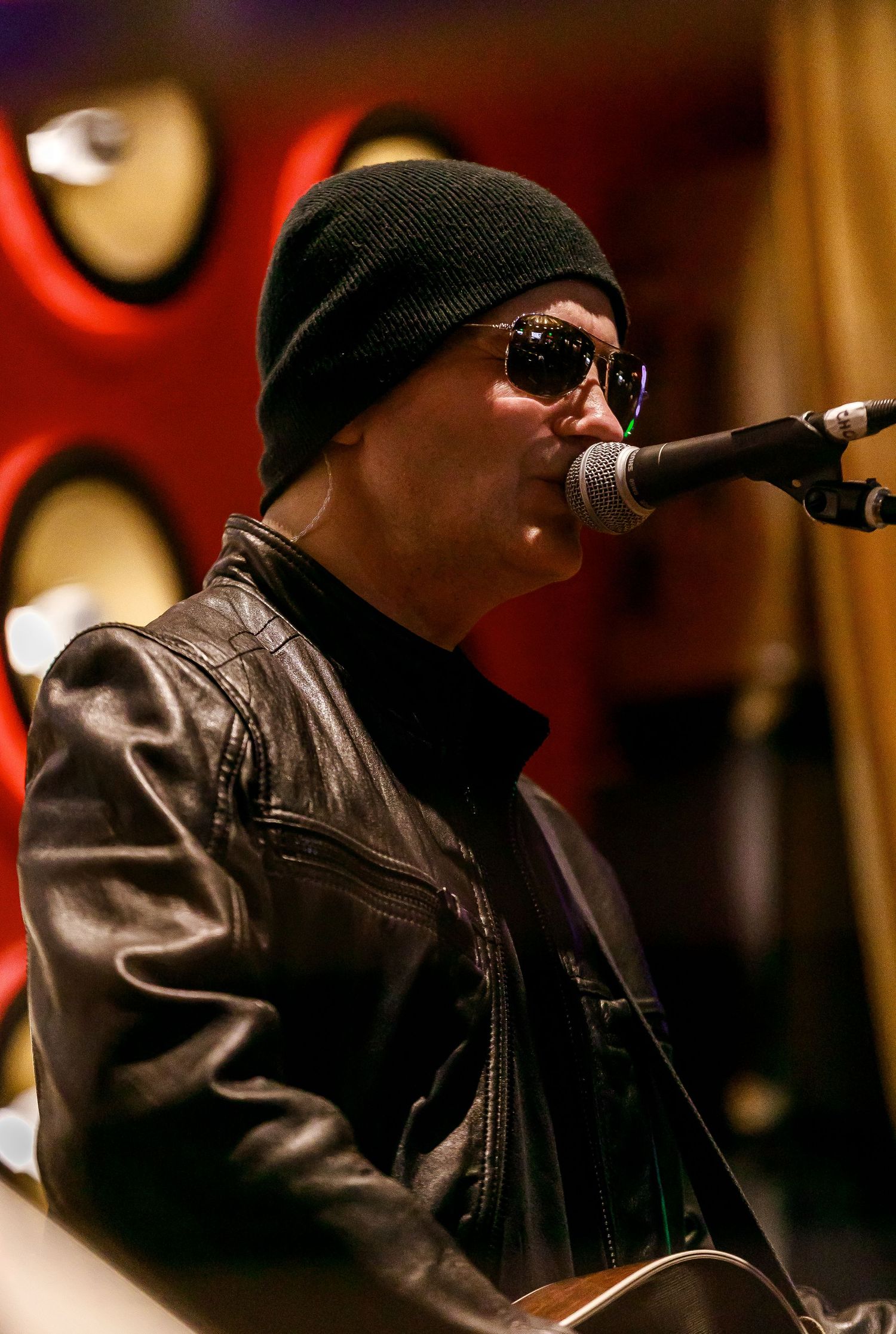 Performer in leather jacket and beanie plays acoustic guitar on stage with dramatic lighting.