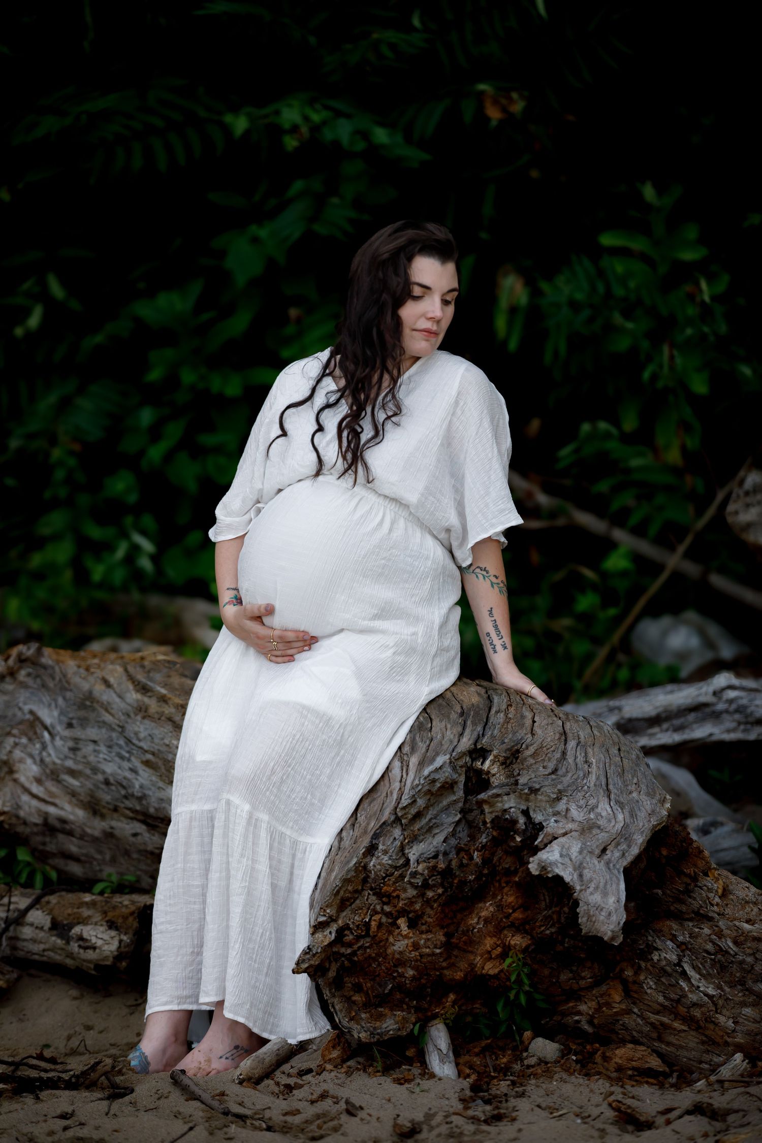 Expectant parent in white dress sits peacefully on driftwood at the beach.