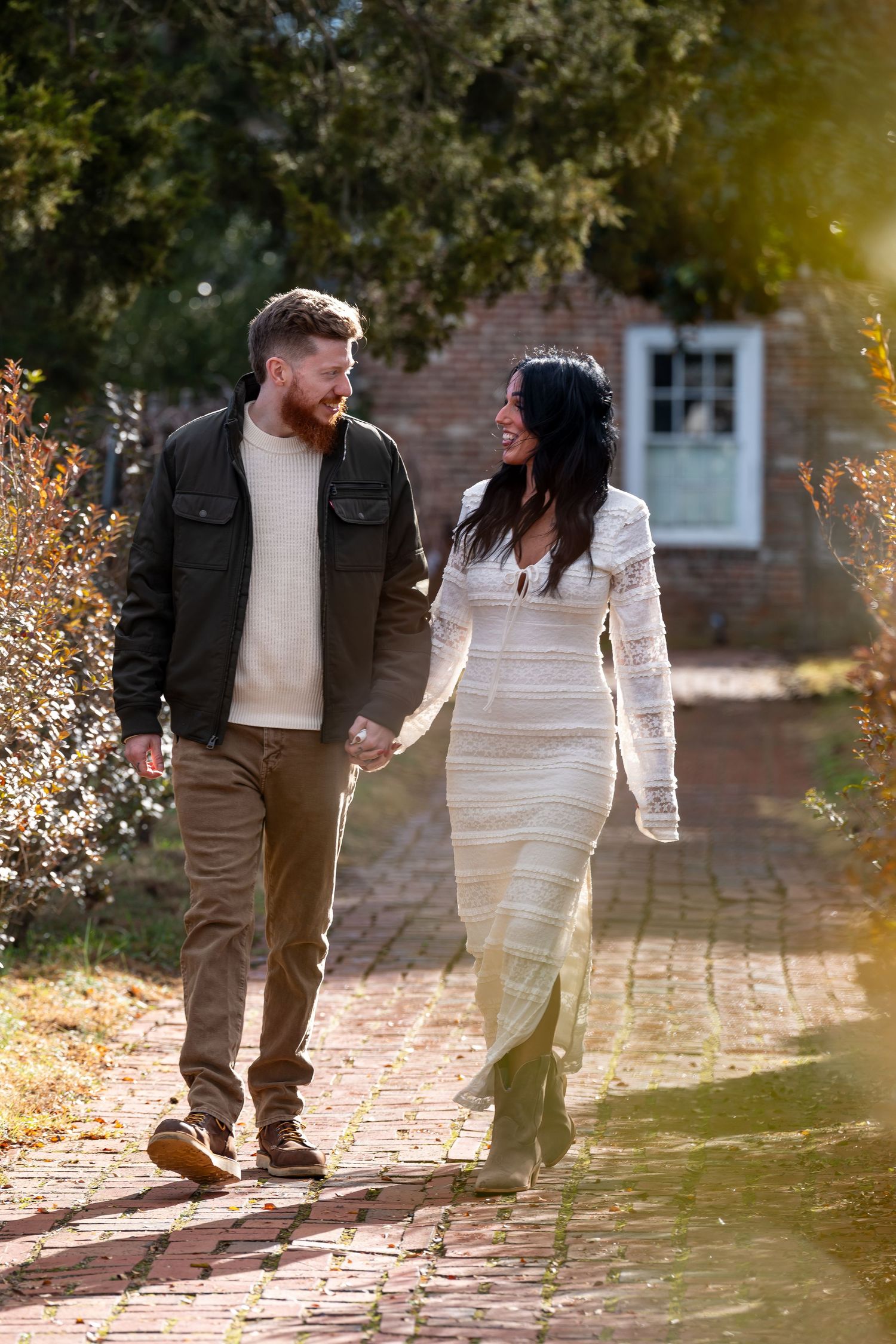 A couple strolls along a brick pathway surrounded by autumn foliage and golden sunlight.