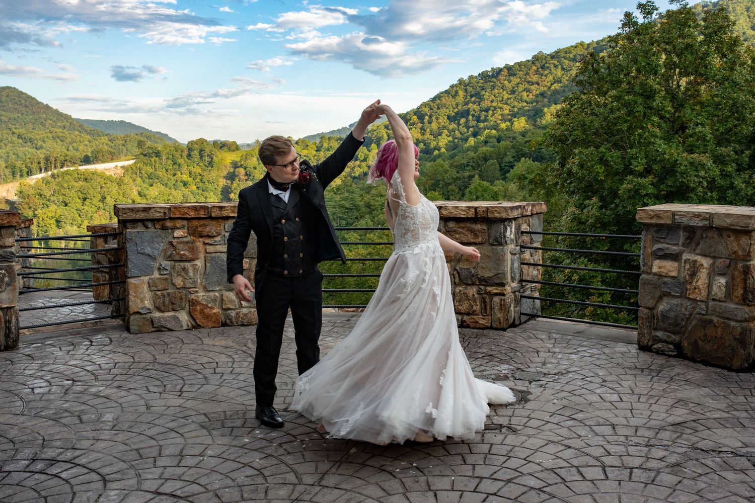 A couple dancing and twirling in formal attire on a stone patio with mountain views in the background.