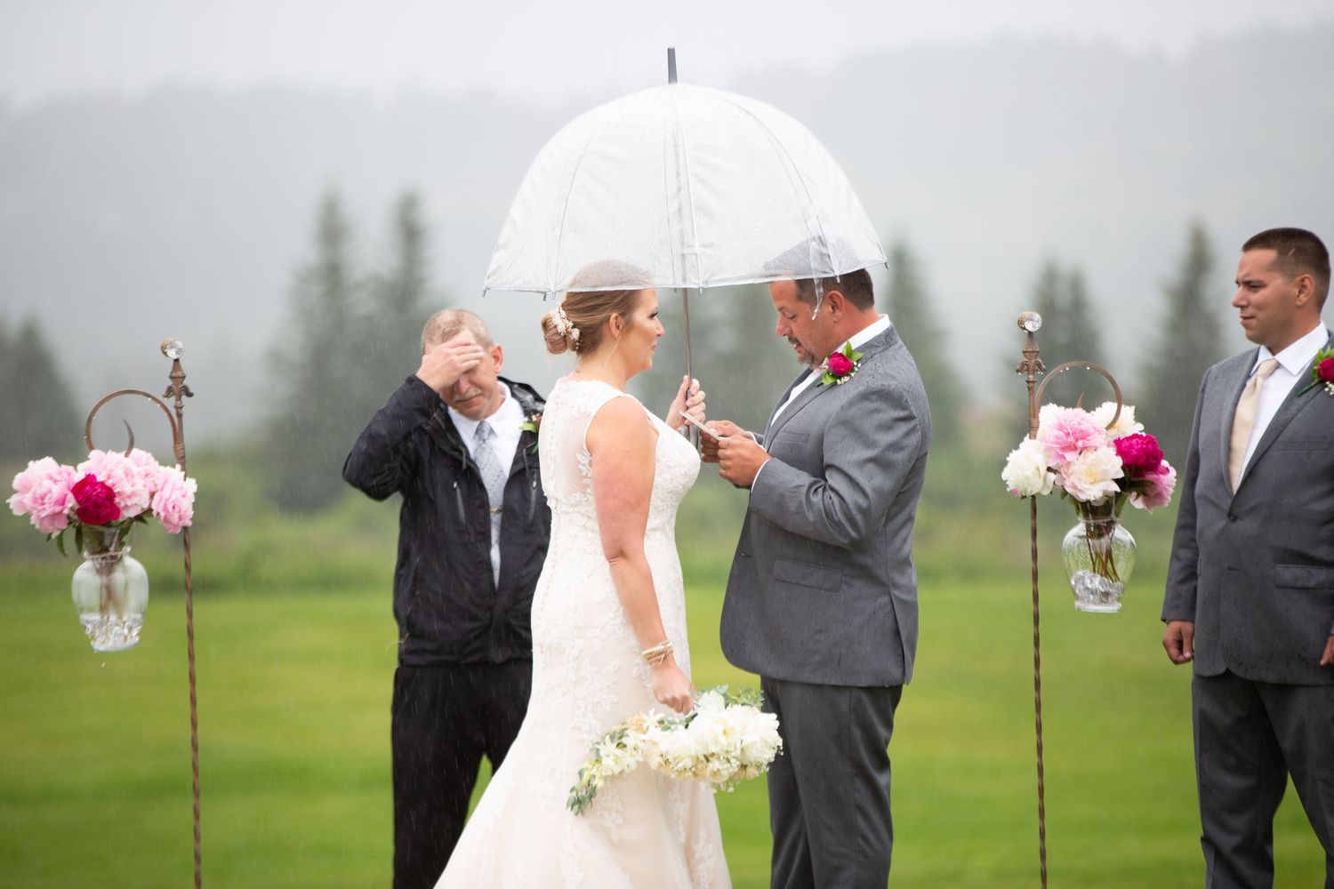 Wedding ceremony outdoors on rainy day with couple under white umbrellas surrounded by wedding party.