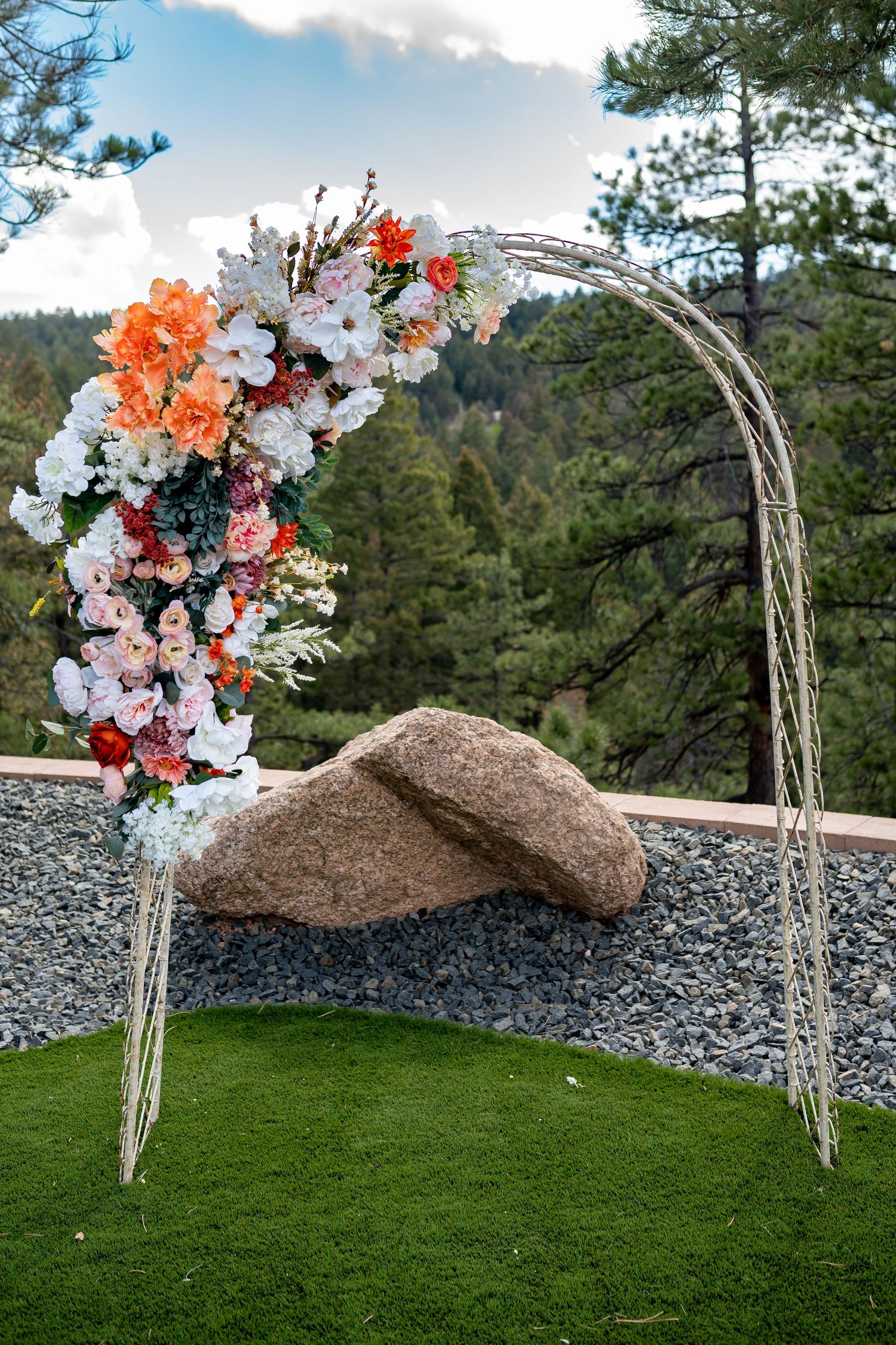 Wedding arch decorated with orange and white flowers against mountain landscape and pine trees.