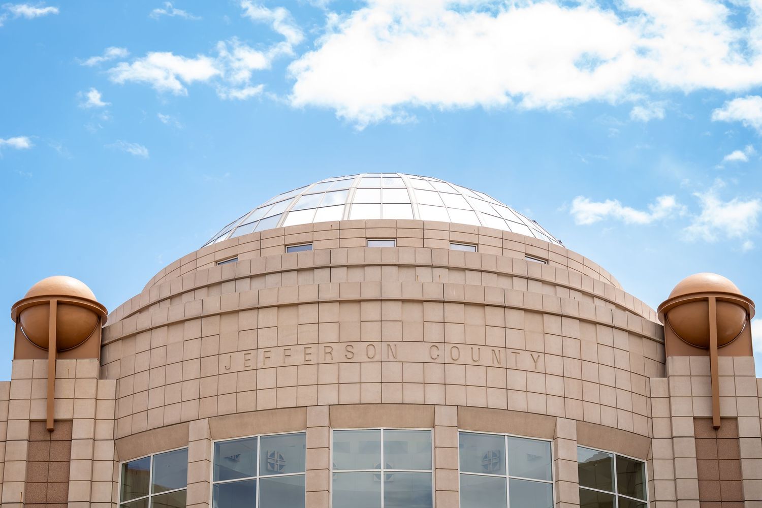 Modern domed building with tan stone exterior against bright blue sky with fluffy white clouds.