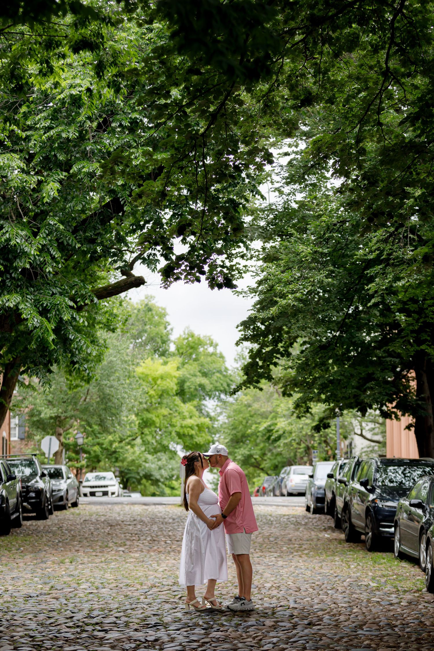 Two people stand together on a tree-lined cobblestone street with parked cars visible in the background.