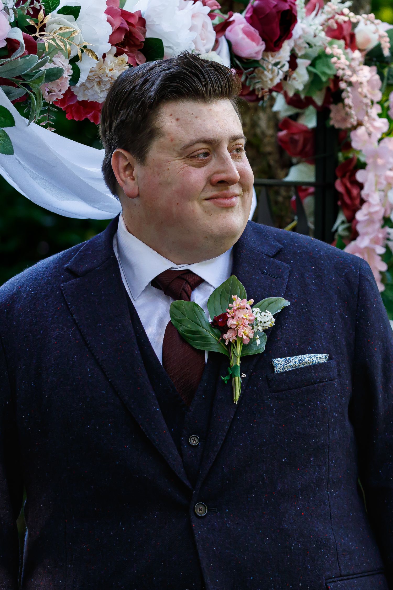 Groom in a navy suit with burgundy tie and boutonniere at an outdoor wedding.