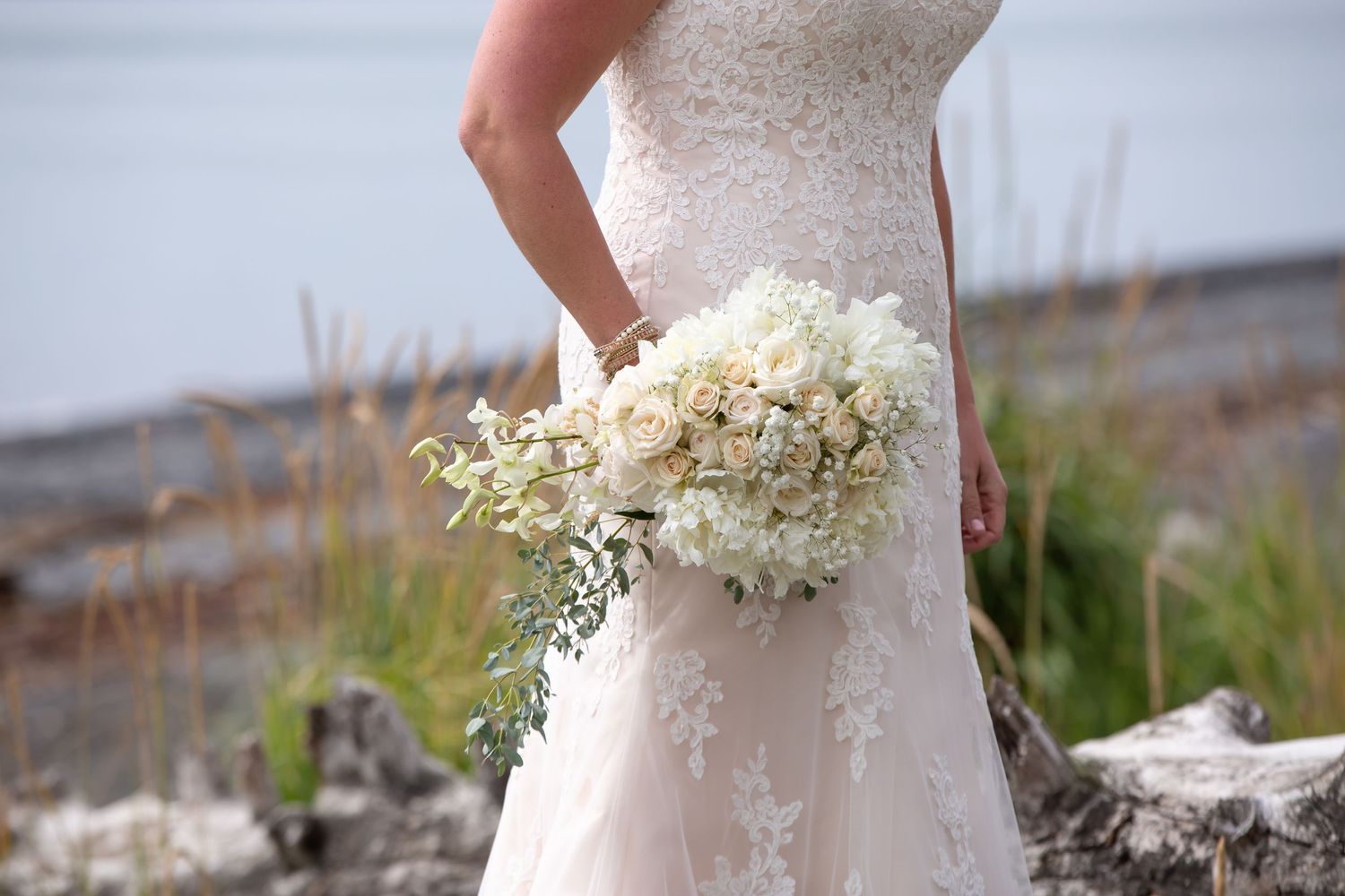 Elegant lace wedding dress detail with floral bouquet against outdoor backdrop.