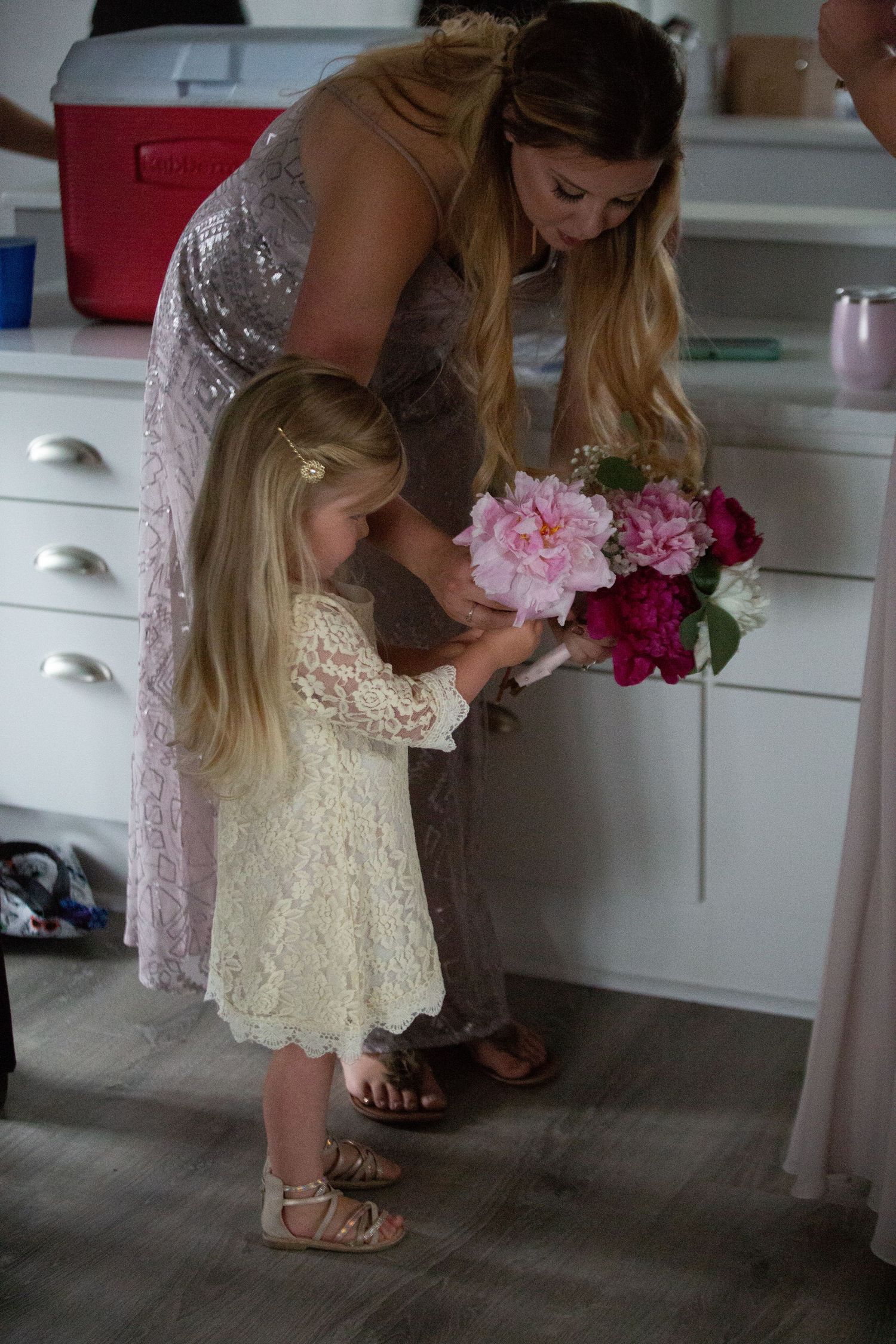 Two people in lace dresses admiring pink and purple flower bouquet in a kitchen.