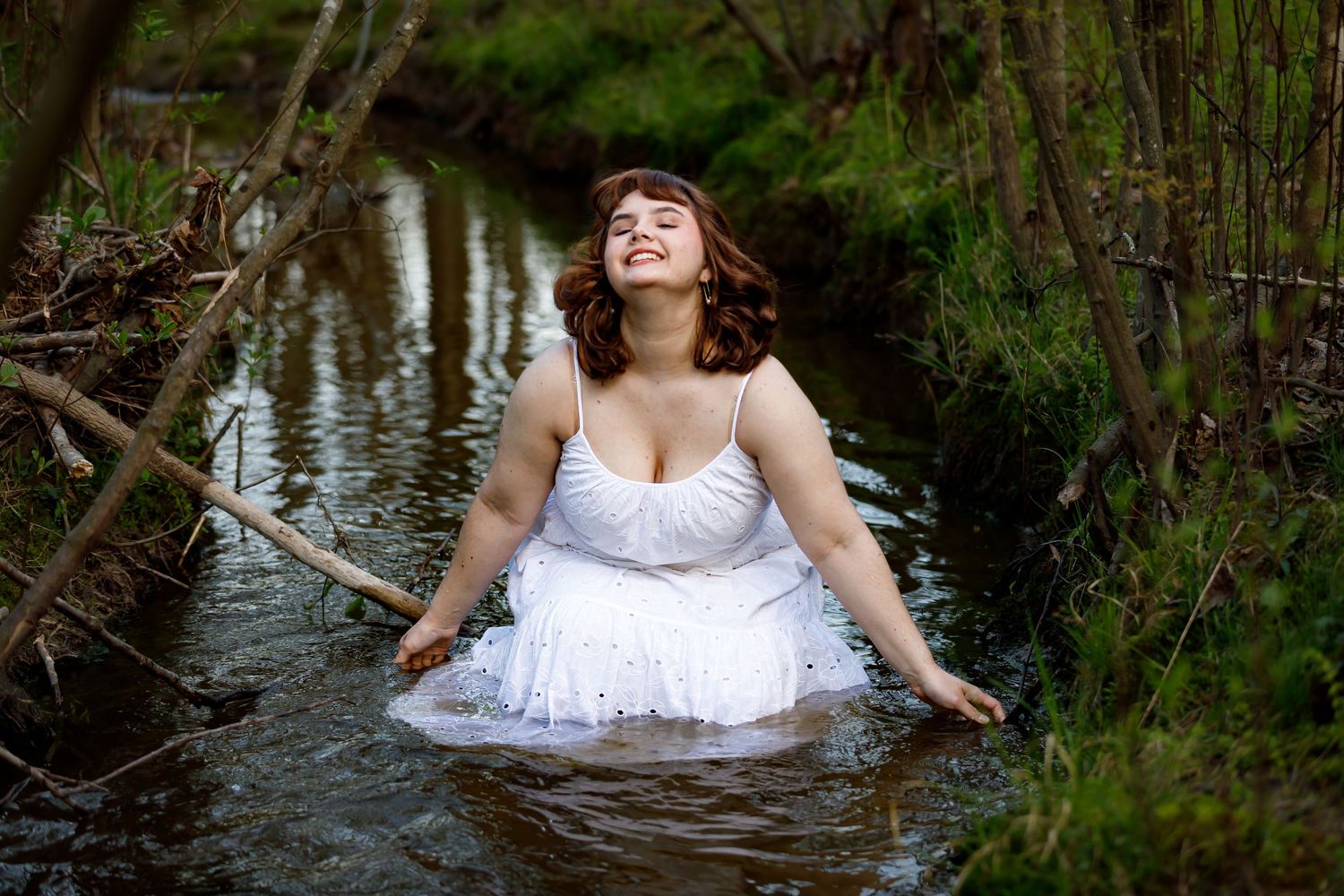 Figure in white dress poses dramatically in a natural water setting surrounded by trees.