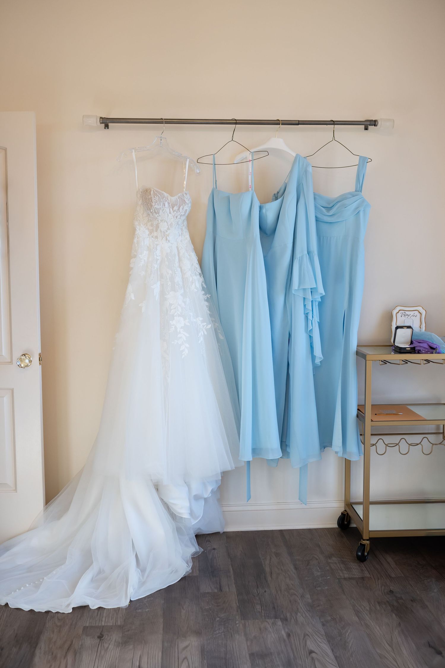 Wedding dress and light blue bridesmaid dresses hanging on display rack against neutral wall.