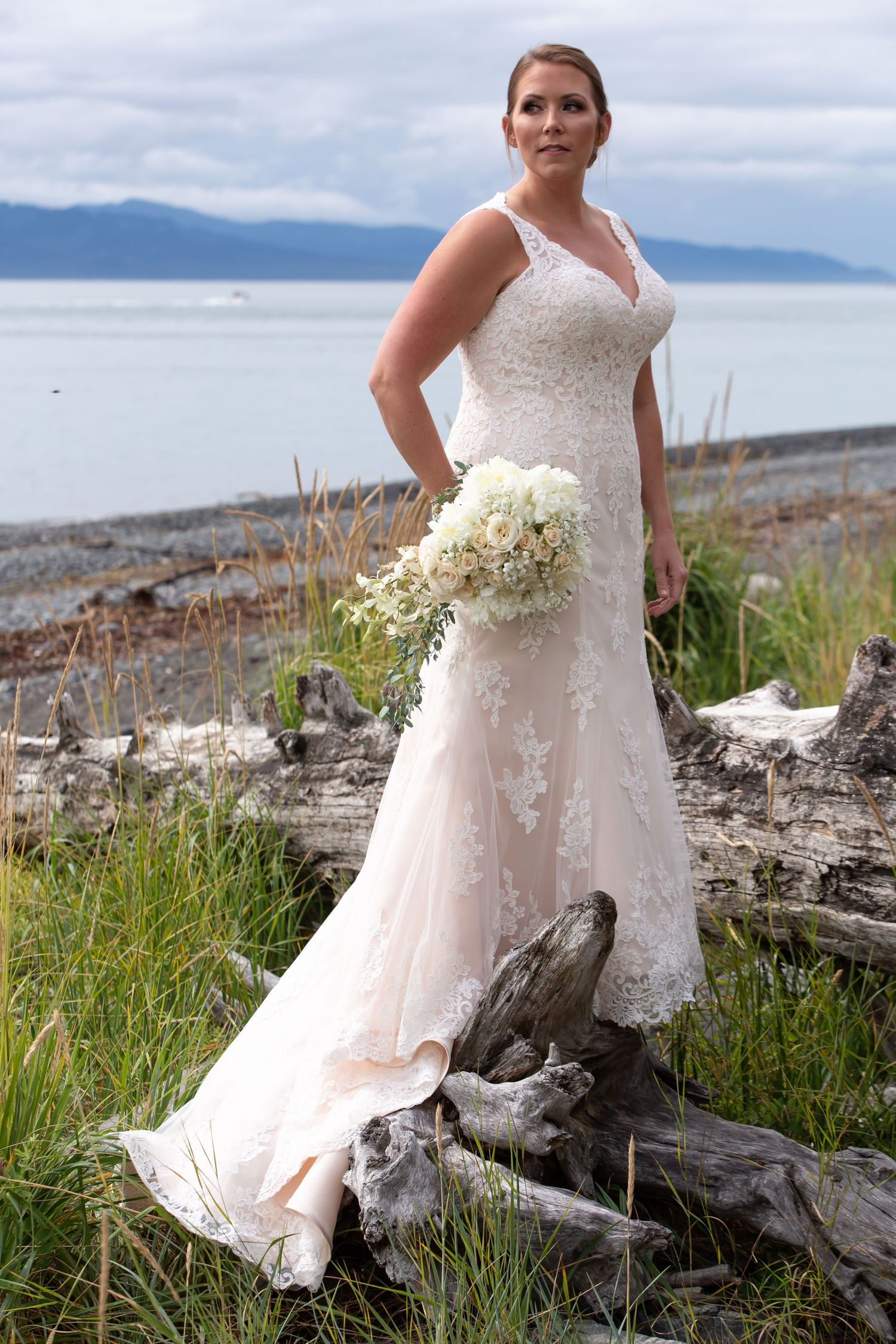 A flowing white wedding dress is showcased against a scenic coastal backdrop with driftwood and beach grass.