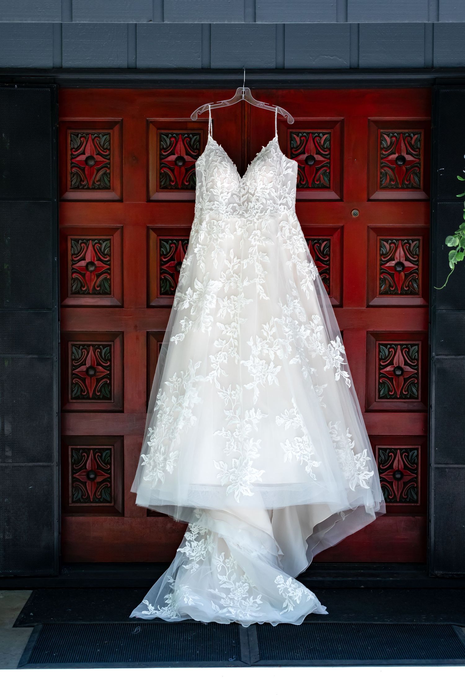 An elegant white lace wedding dress hangs against ornate red wooden doors.