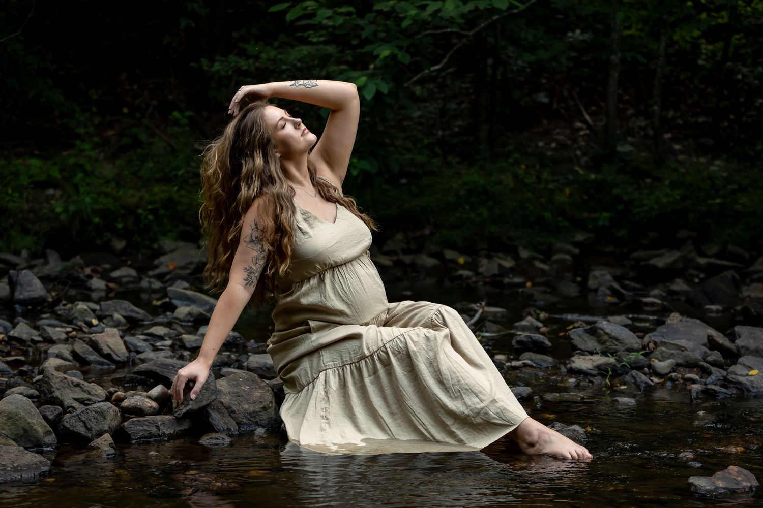 Woman in cream dress sits peacefully by rocky stream in natural setting.