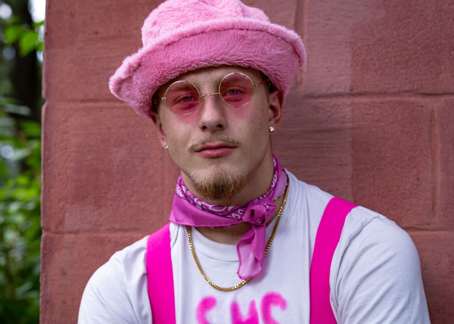 Portrait against brick wall featuring pink hat and bandana with white shirt.