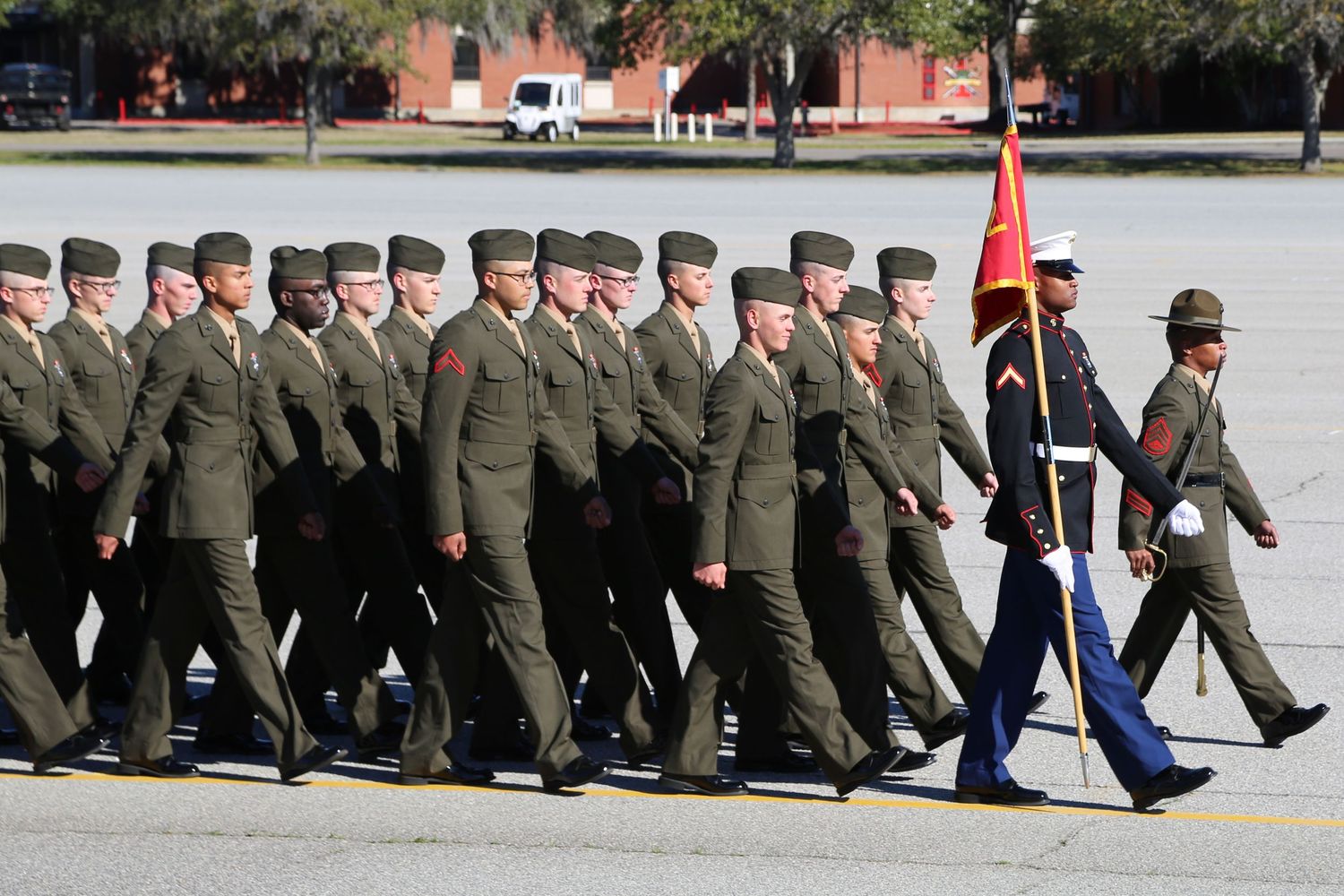 Marine recruits marching in formation led by color guard on parade ground.