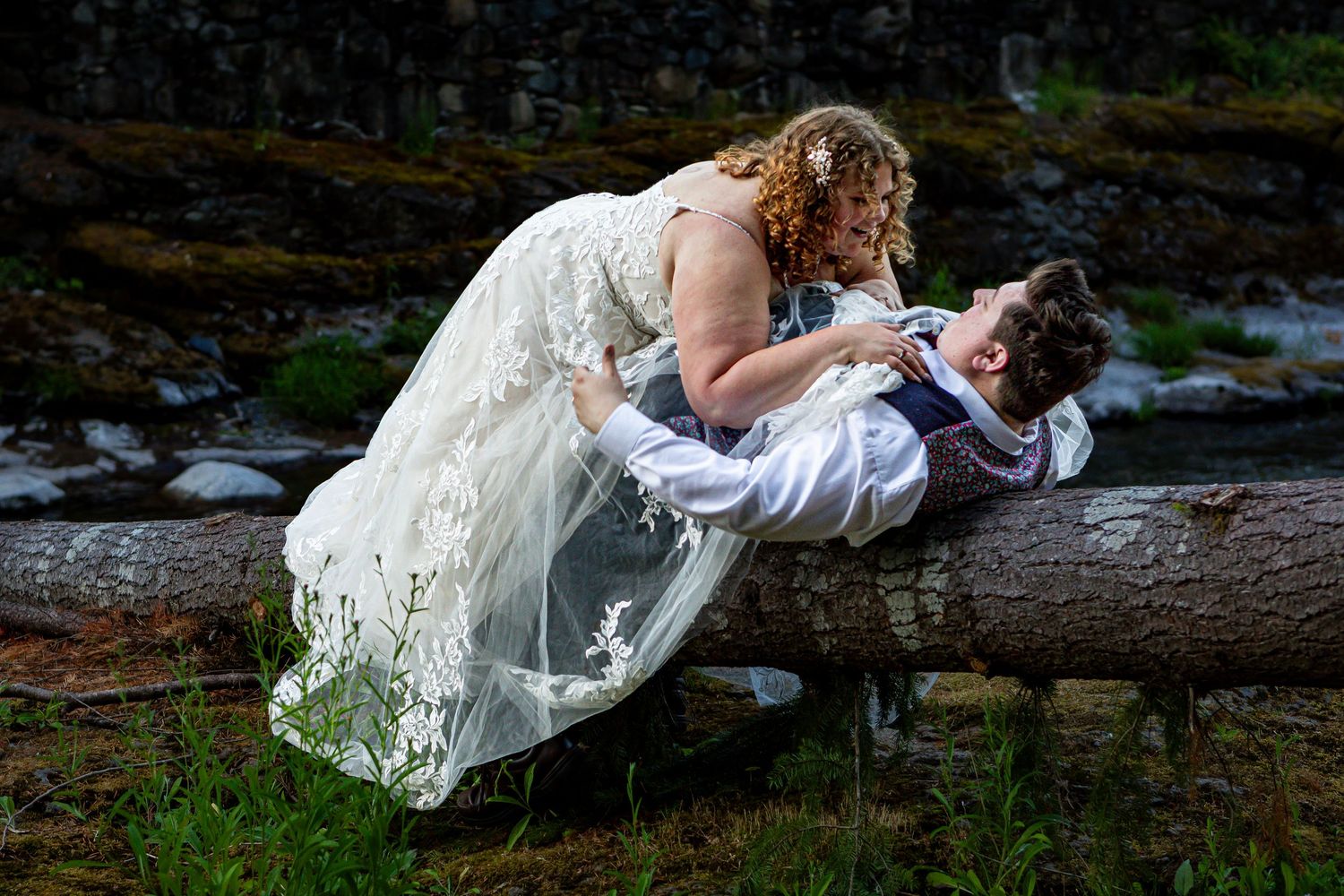 A playful wedding moment captured on a fallen log by a forest stream.
