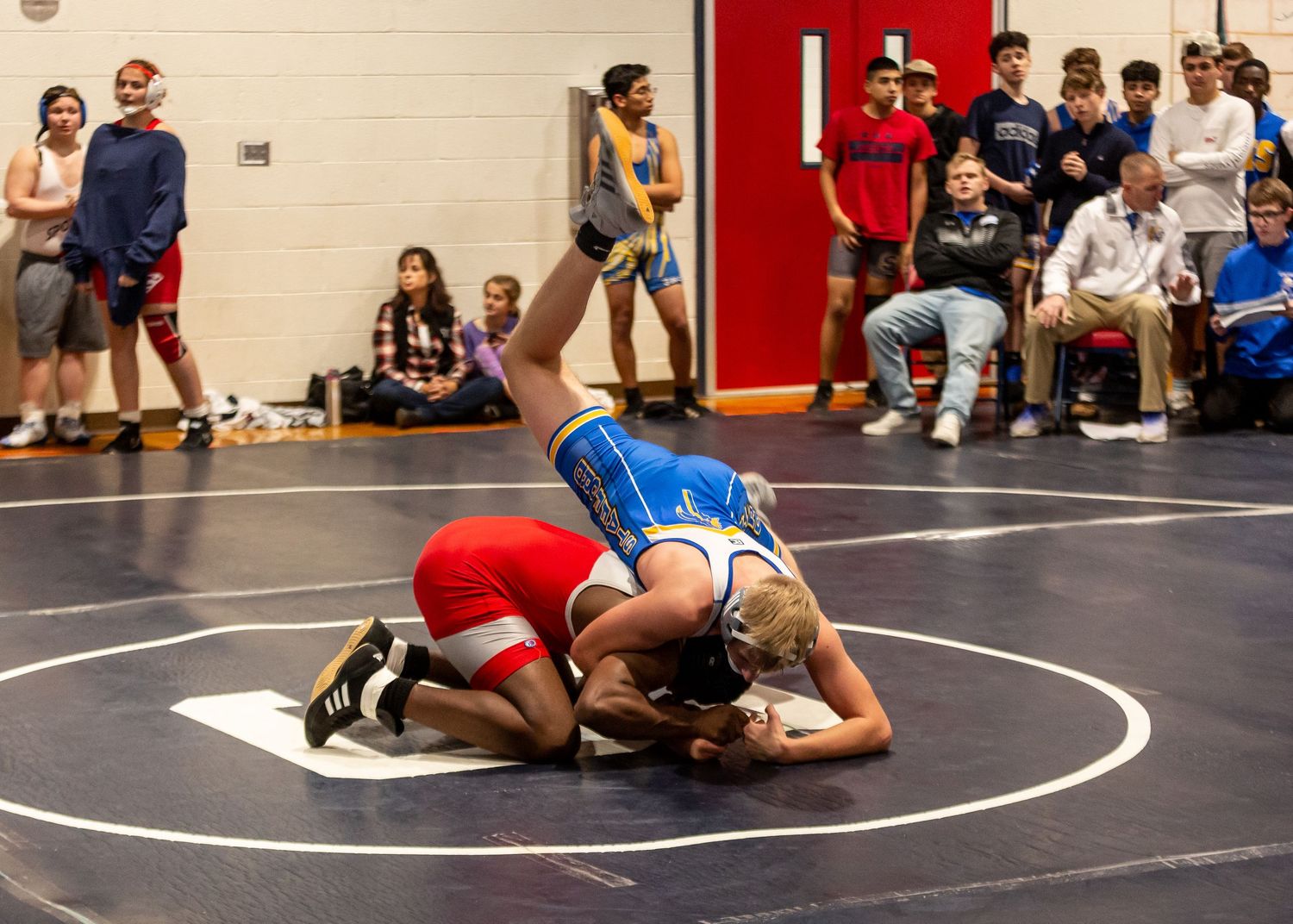 Indoor wrestling match with athletes competing on black mat while crowd watches.