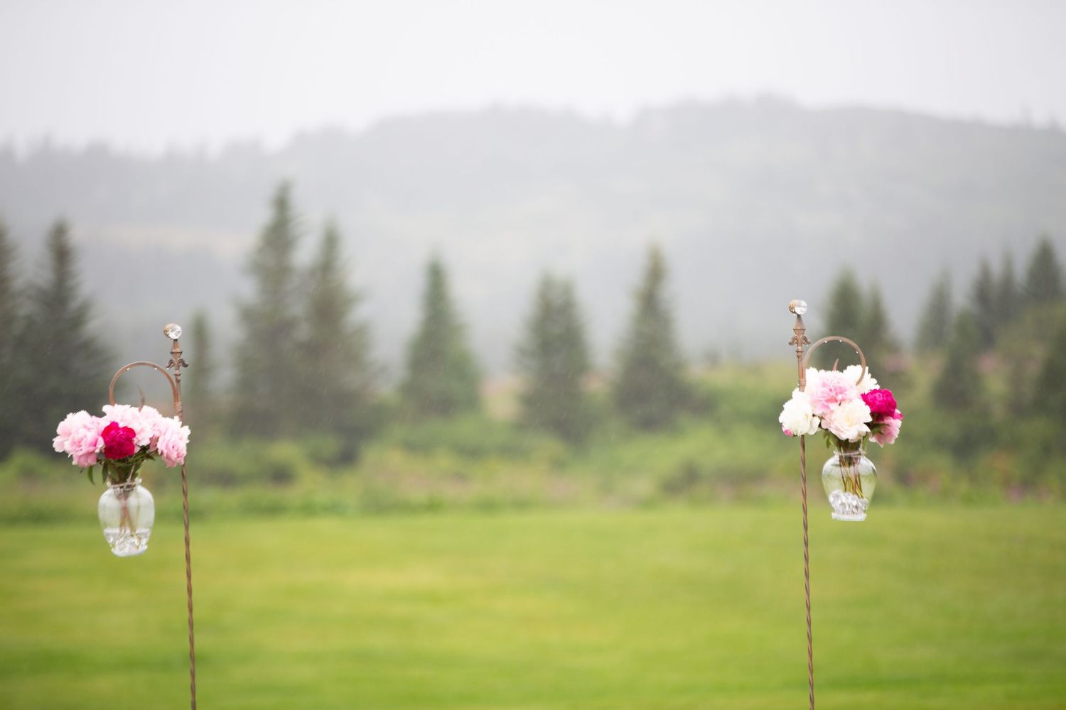 Decorative hanging glass jars with flowers line an outdoor wedding aisle against a misty mountain backdrop.