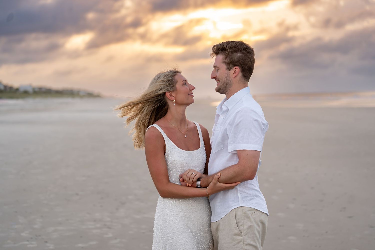 Couple in white outfits embracing on a beach during sunset.
