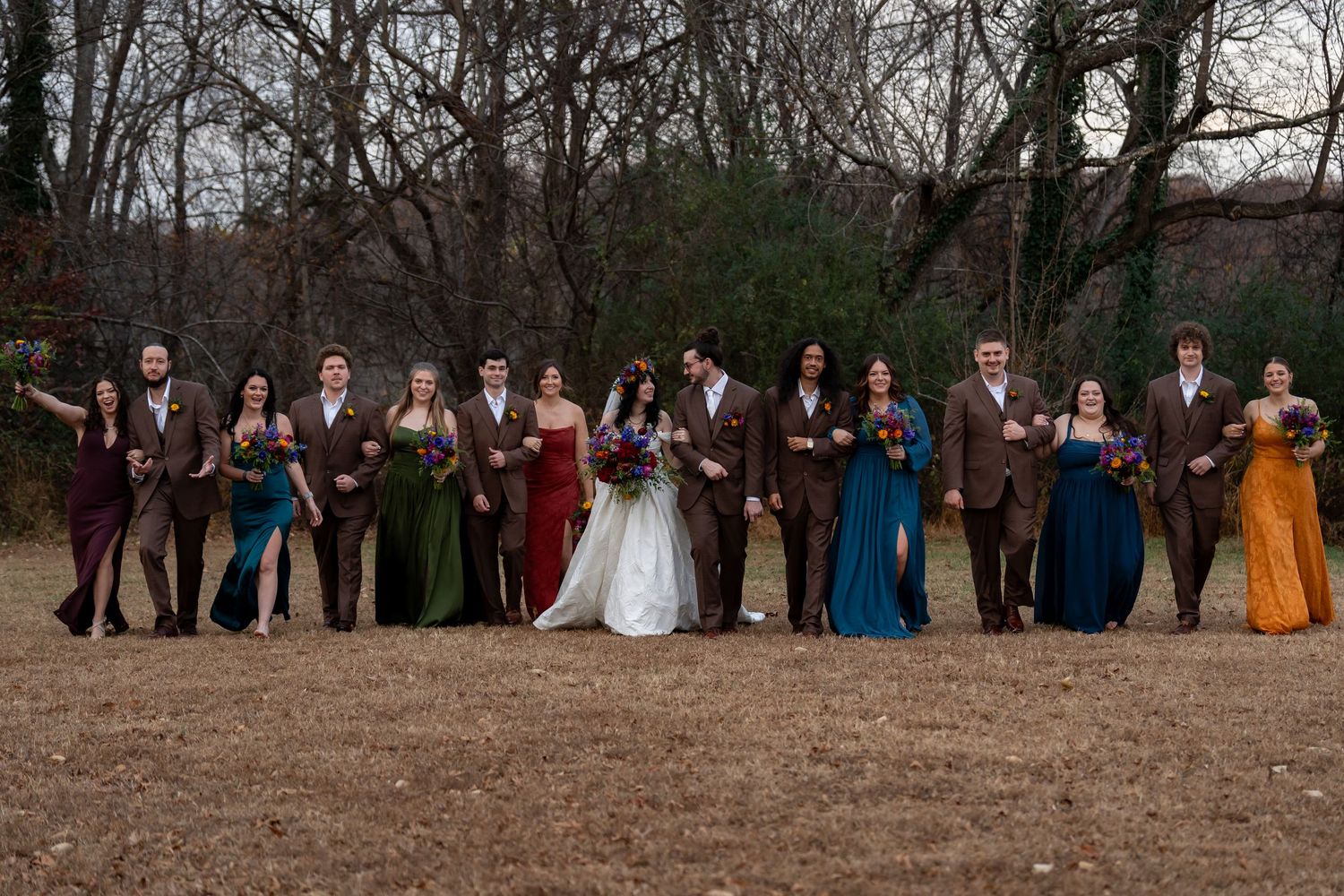 Wedding guests in colorful attire stand in rows during an outdoor ceremony.