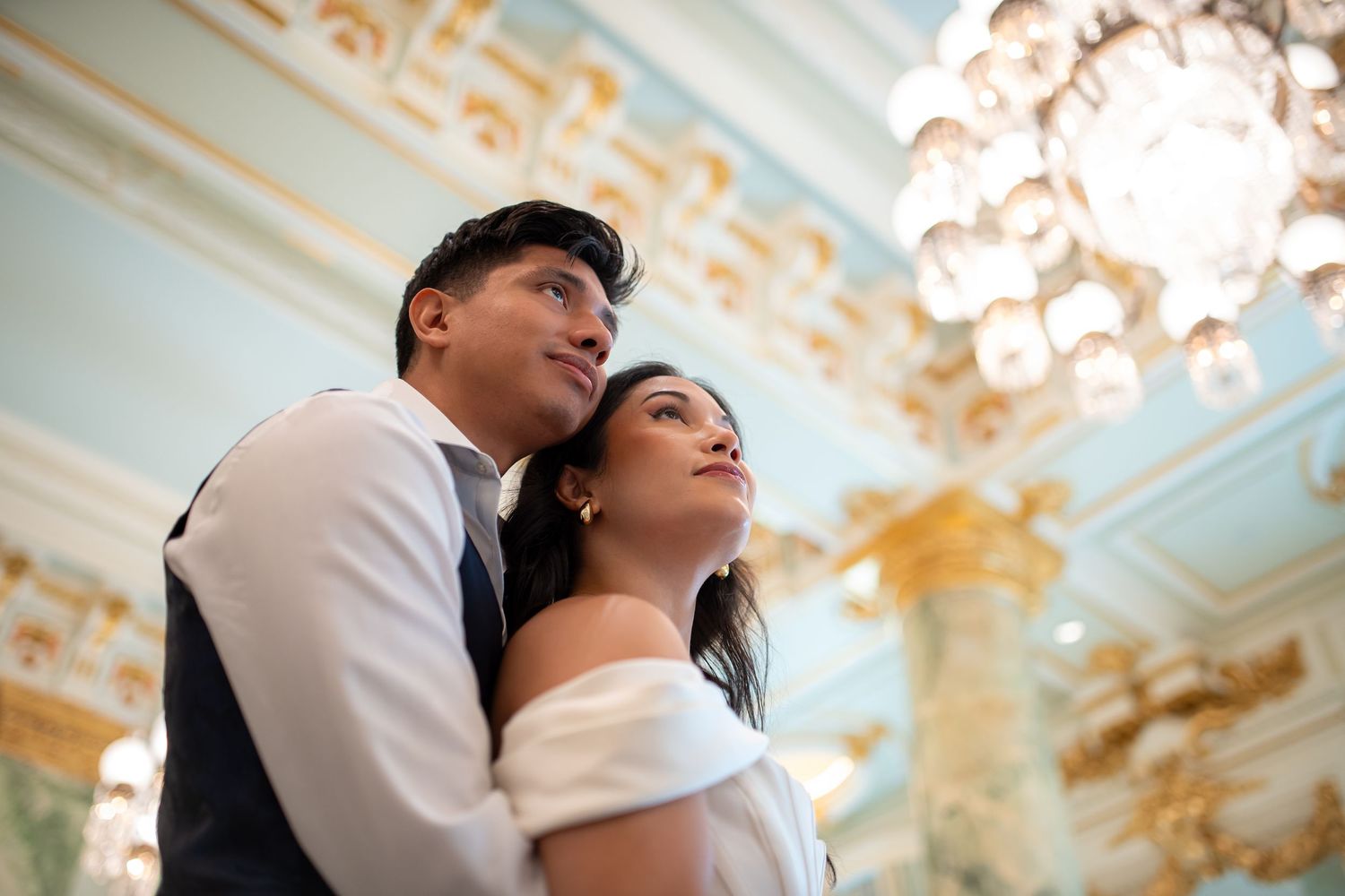 Romantic couple poses under an ornate chandelier in an elegant ballroom setting.