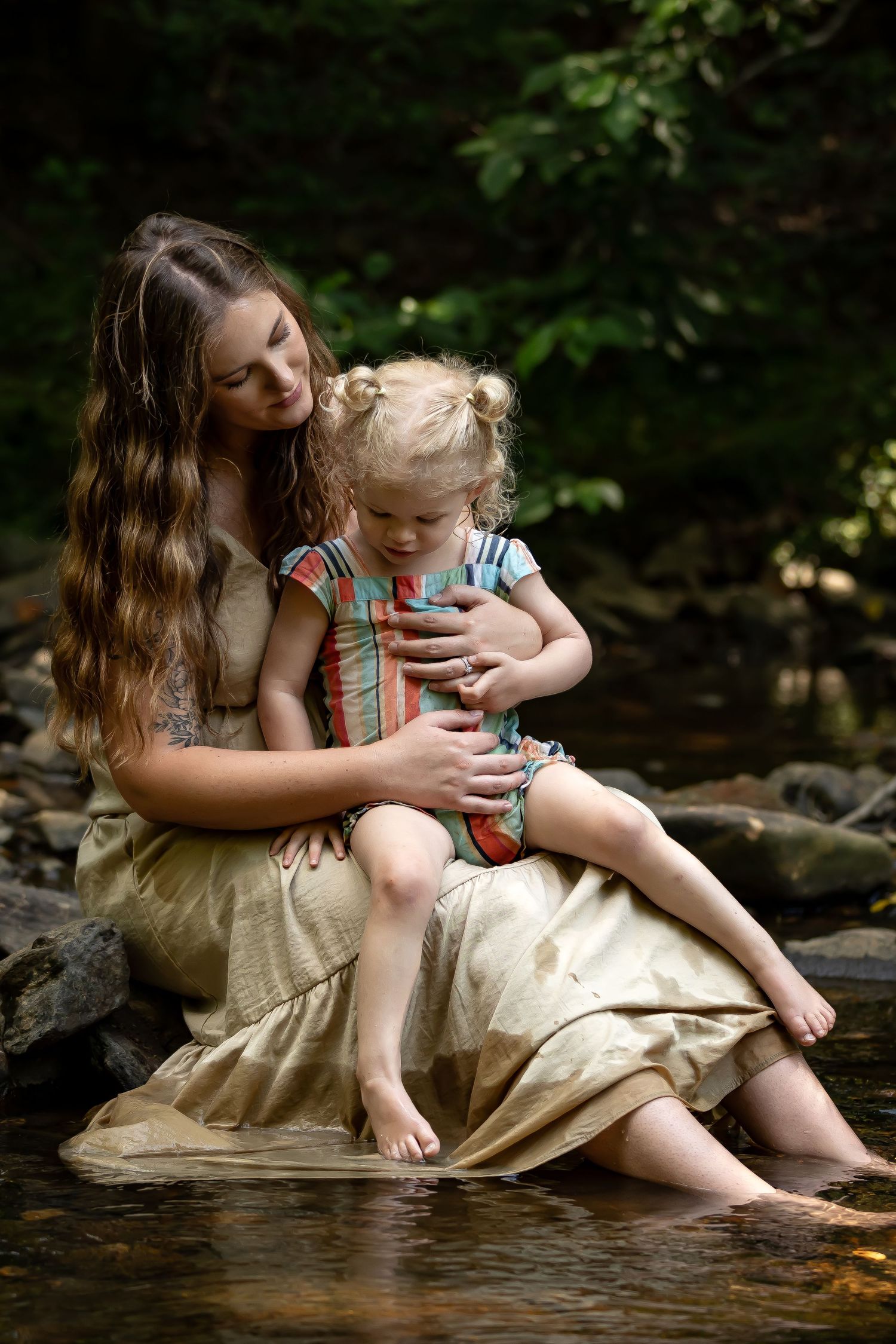 A tender moment shared between parent and child sitting by a stream in a forest setting.