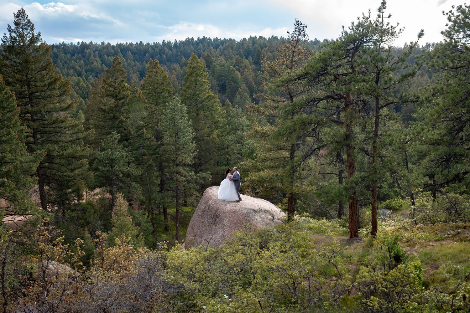 Bride and groom with bride in white wedding dress on a large boulder surrounded by evergreen forest in the Rocky mountains in Colorado.