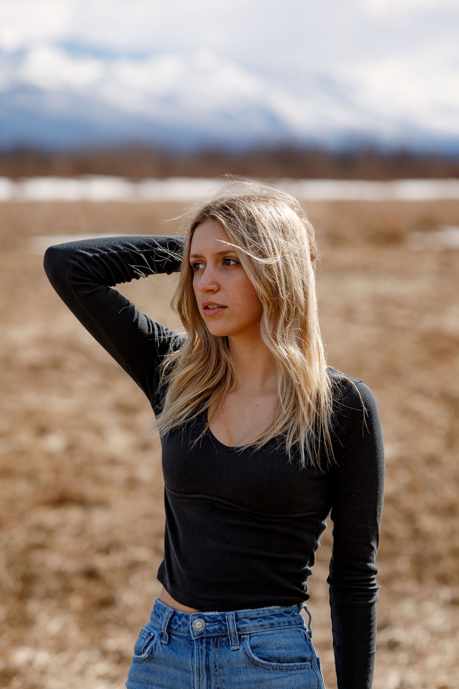 A person in a black long-sleeve top and jeans poses in a field with mountains in the background.