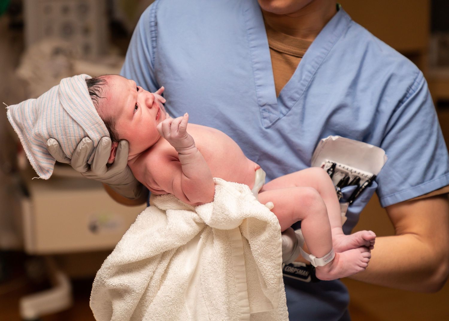 Healthcare worker in blue scrubs holding and caring for newborn baby wrapped in cream blanket.