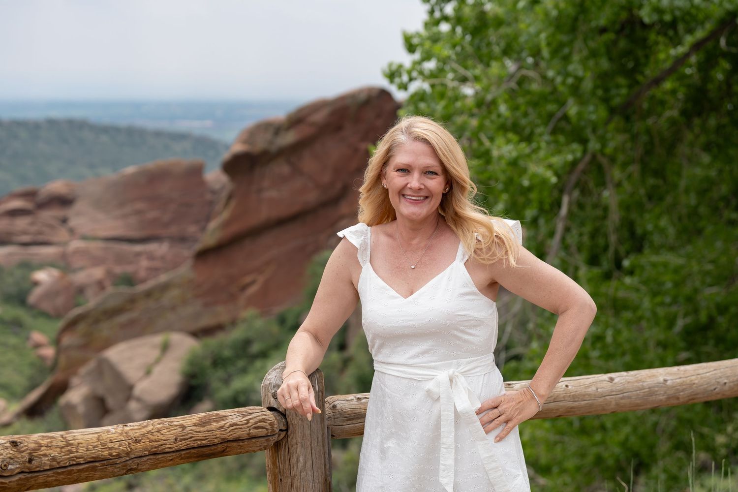 Woman in white sundress poses by wooden railing with red rocks in background.