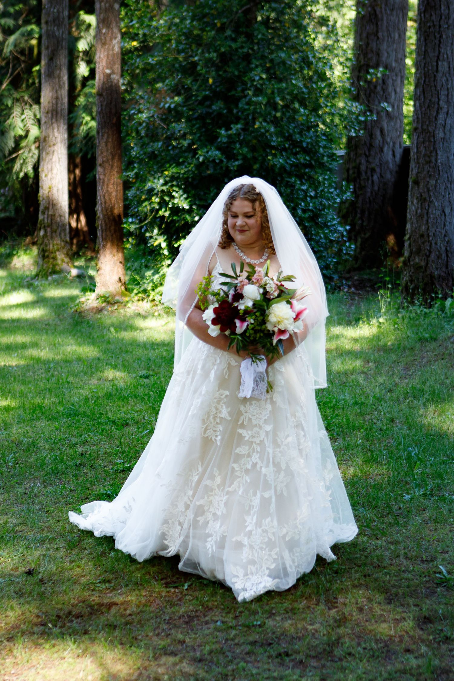 A bride in a flowing white lace gown poses among pine trees in an outdoor setting.