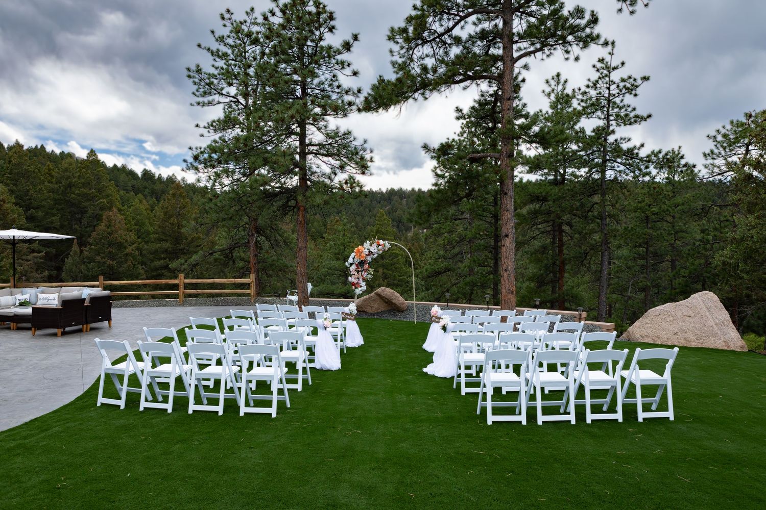 Outdoor wedding ceremony setup with white chairs on grass surrounded by pine trees.