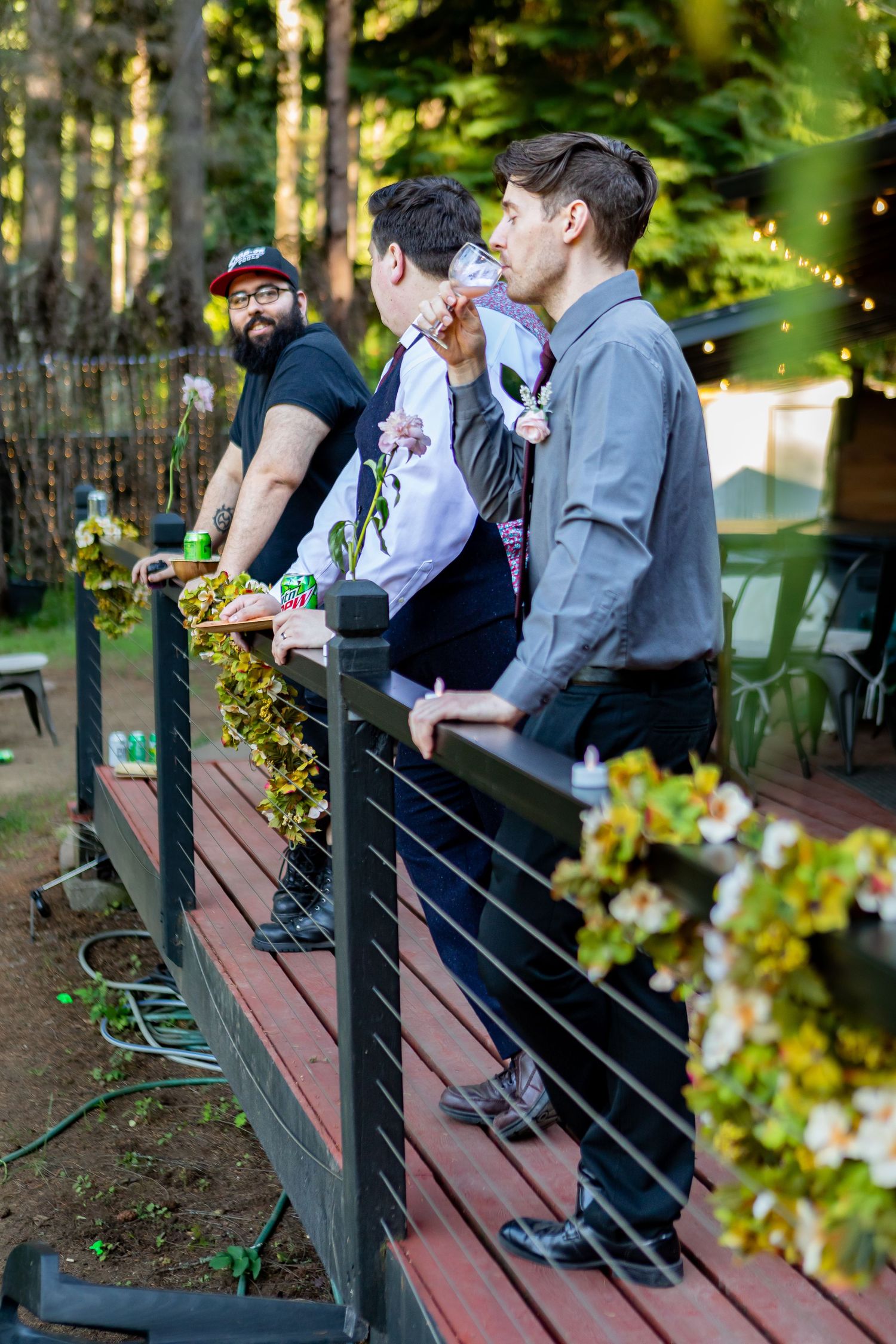 Wedding guests sit on wooden benches in a garden with string lights overhead.