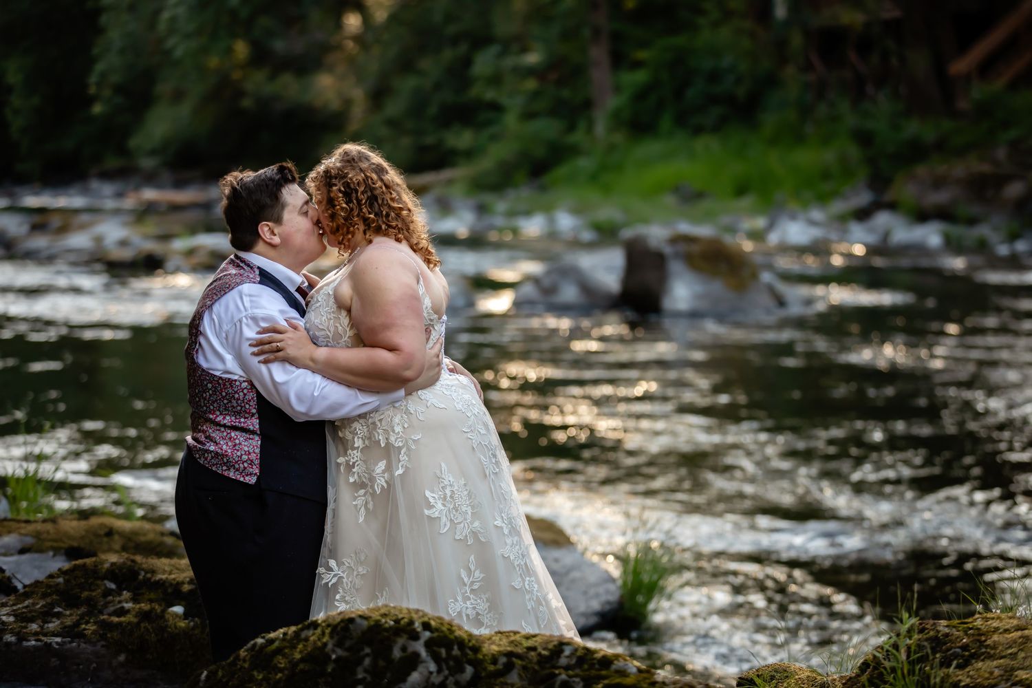 Couple embracing by rocky riverbank during wedding photoshoot.