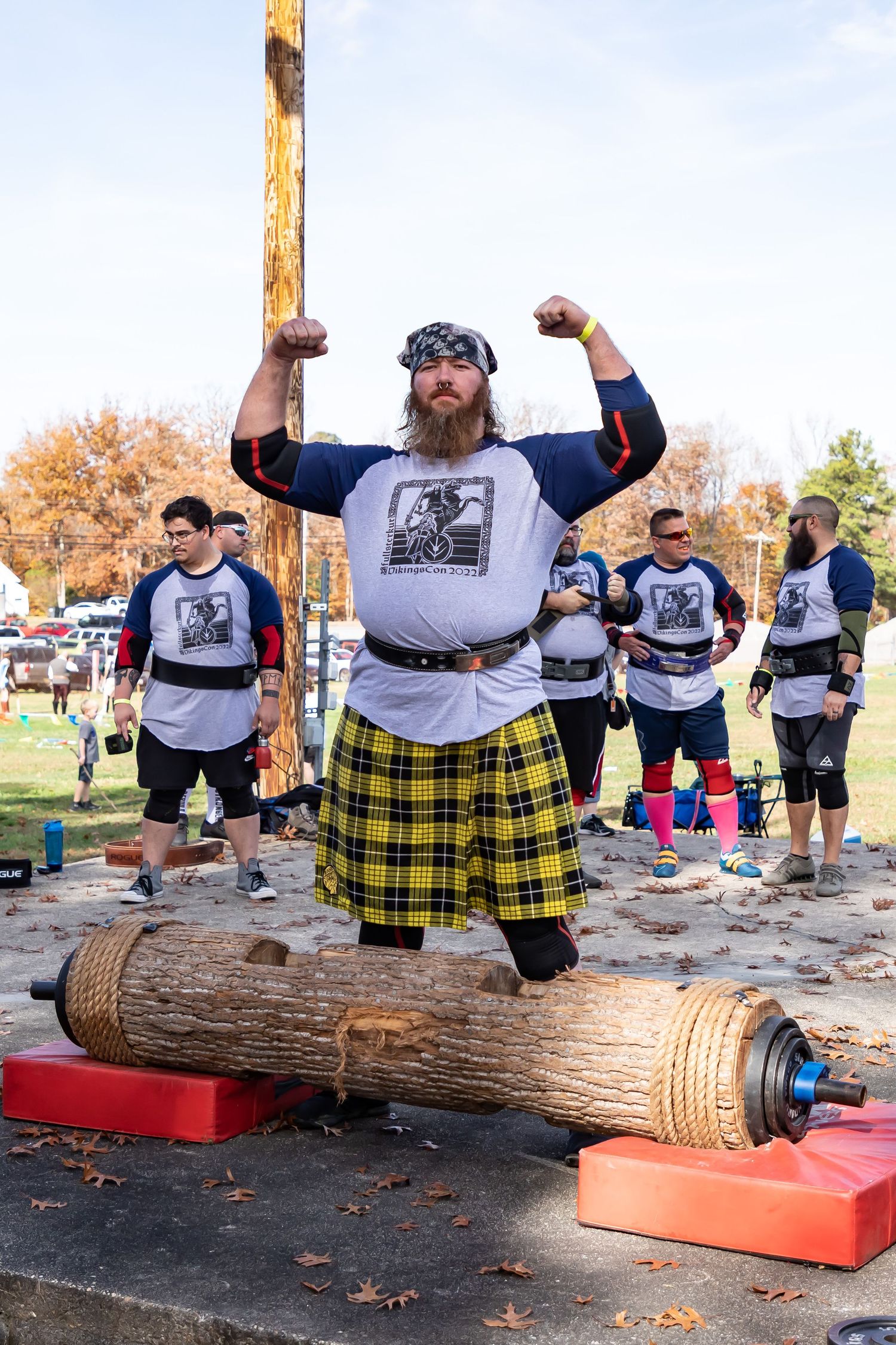 Competitor in plaid kilt attempts heavy log lift during outdoor strongman competition.