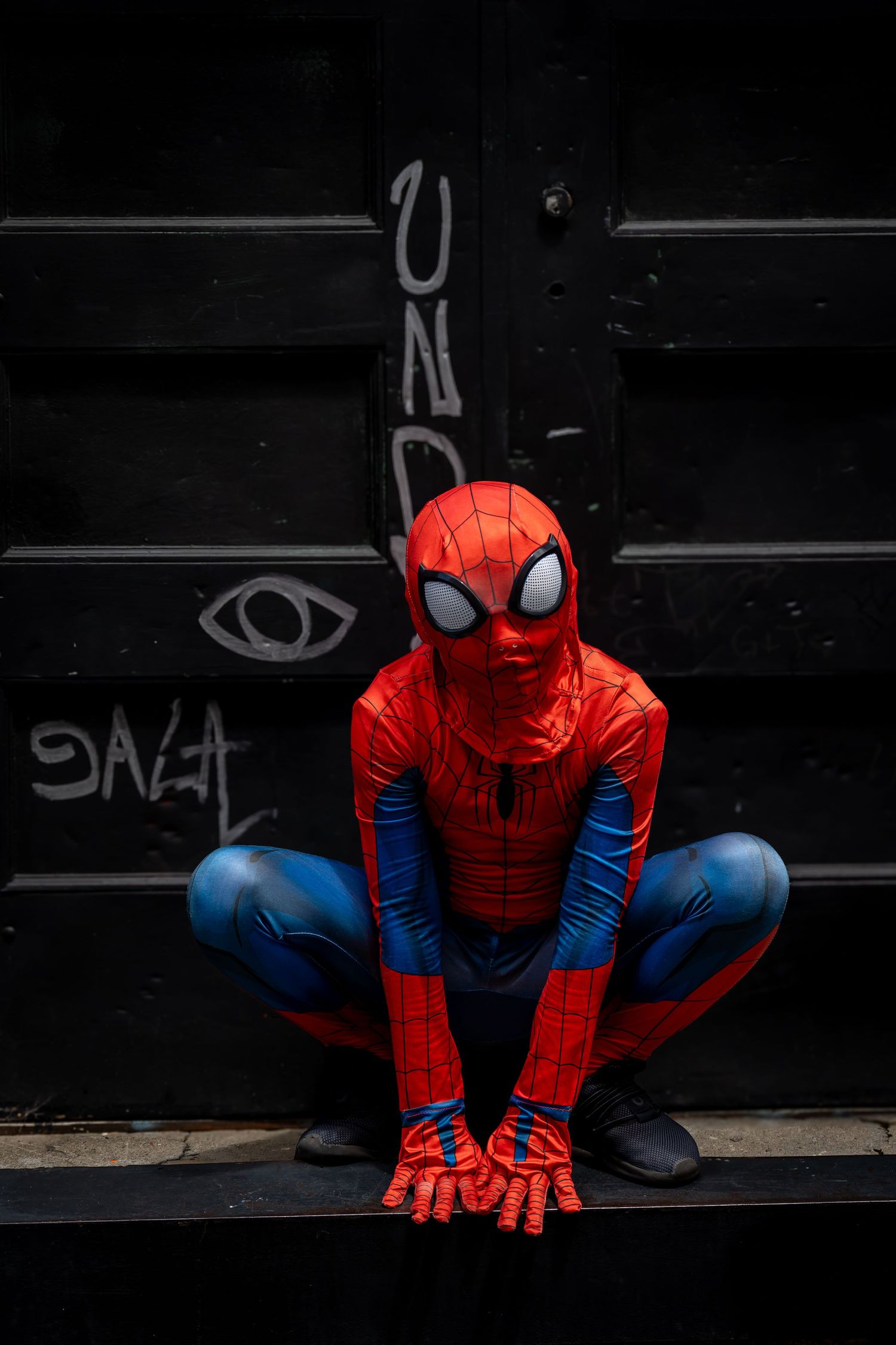 Spider-Man costume wearer crouching in signature pose against dark background with brick wall.