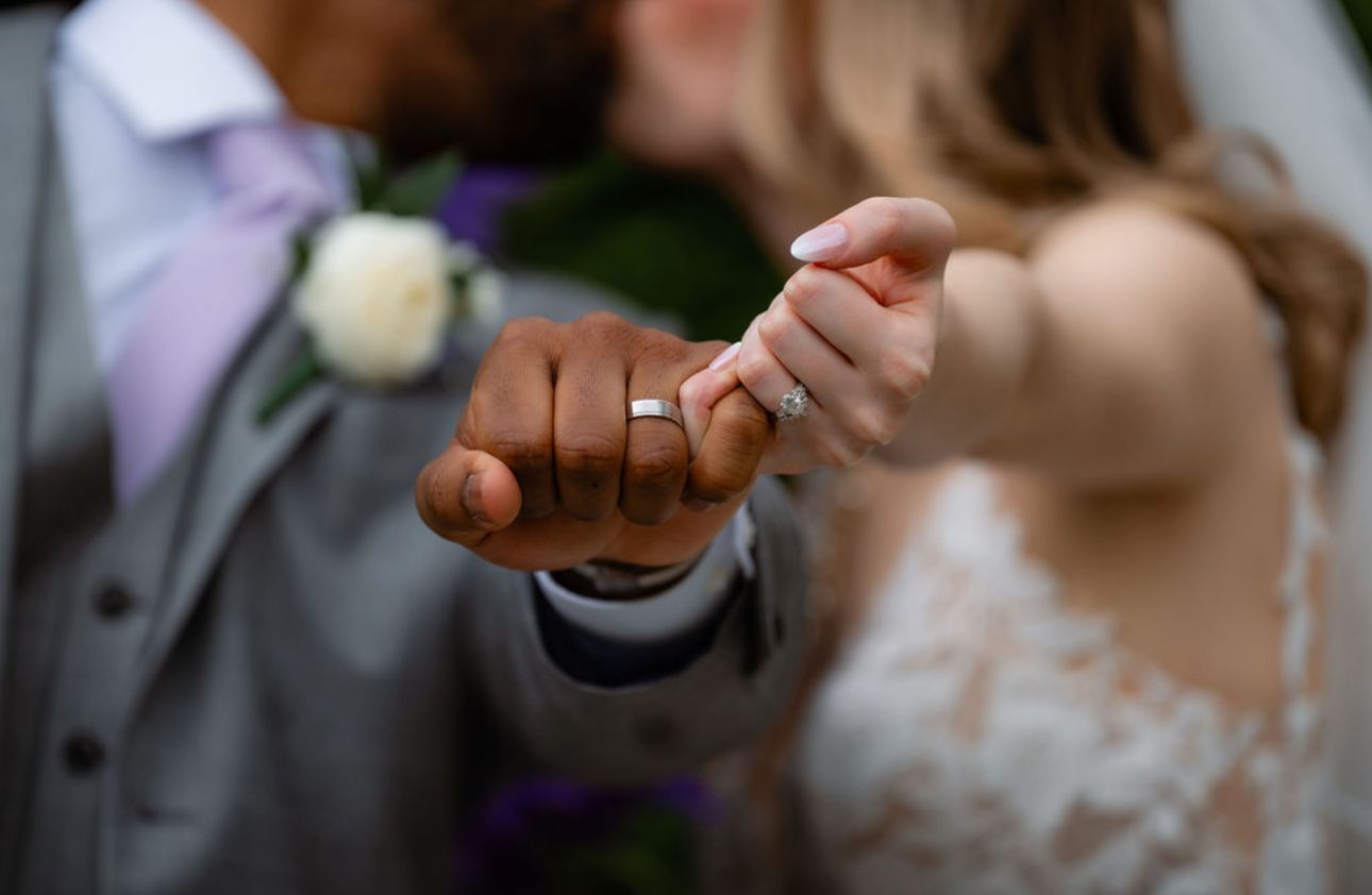 A couple holds hands during their wedding ceremony while exchanging rings with guests watching in the background.
