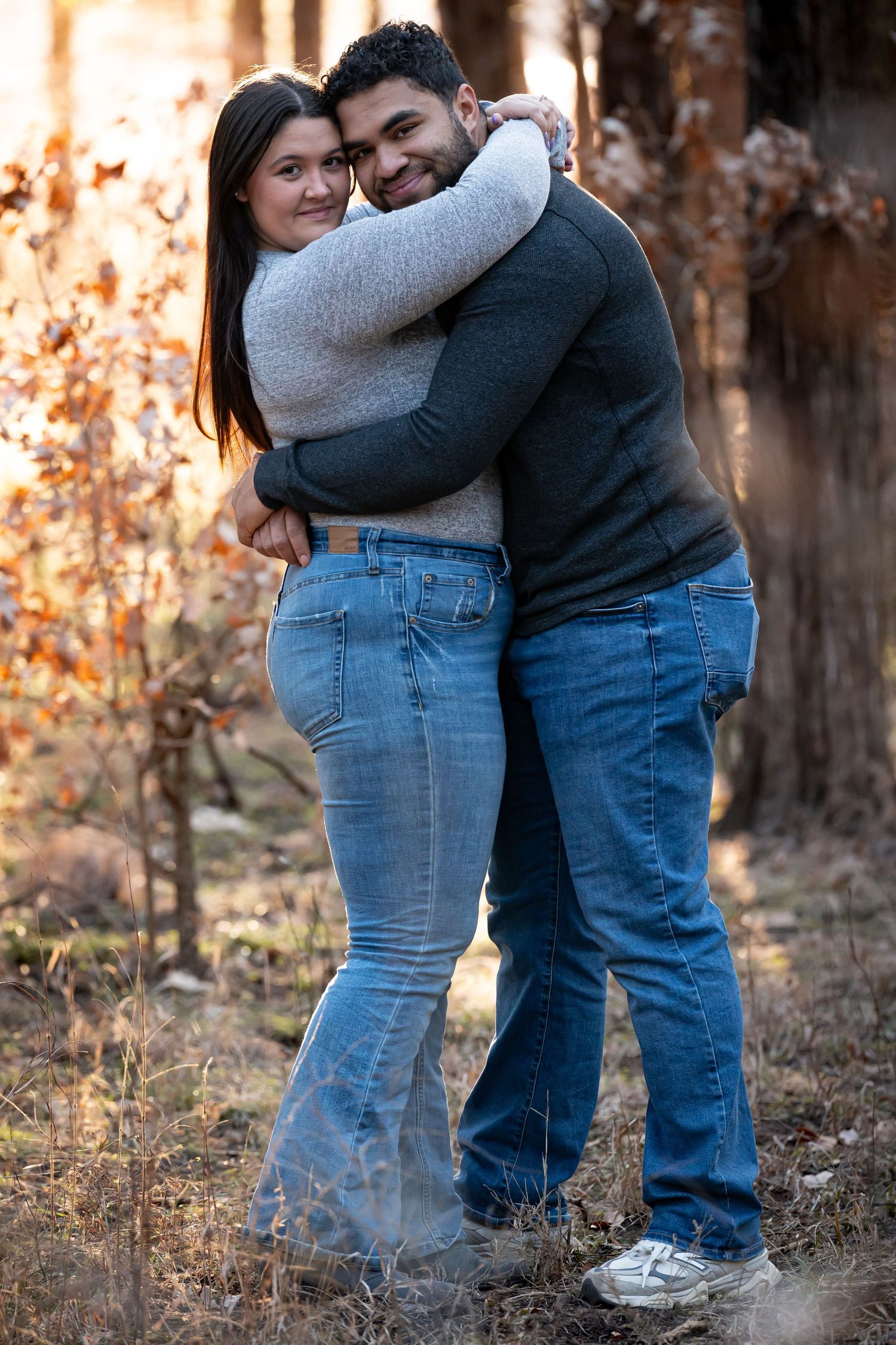 A couple embraces in casual denim and sweaters during an autumn outdoor photoshoot with warm lighting.
