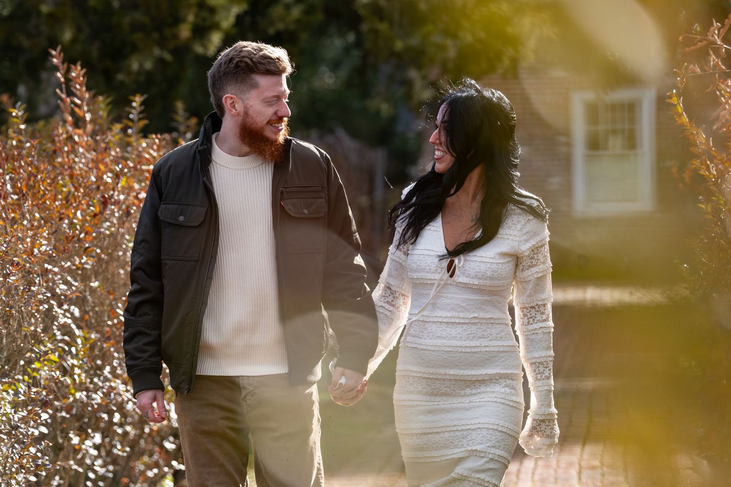 A couple walks together on an autumn path with fallen leaves and warm sunlight filtering through trees.