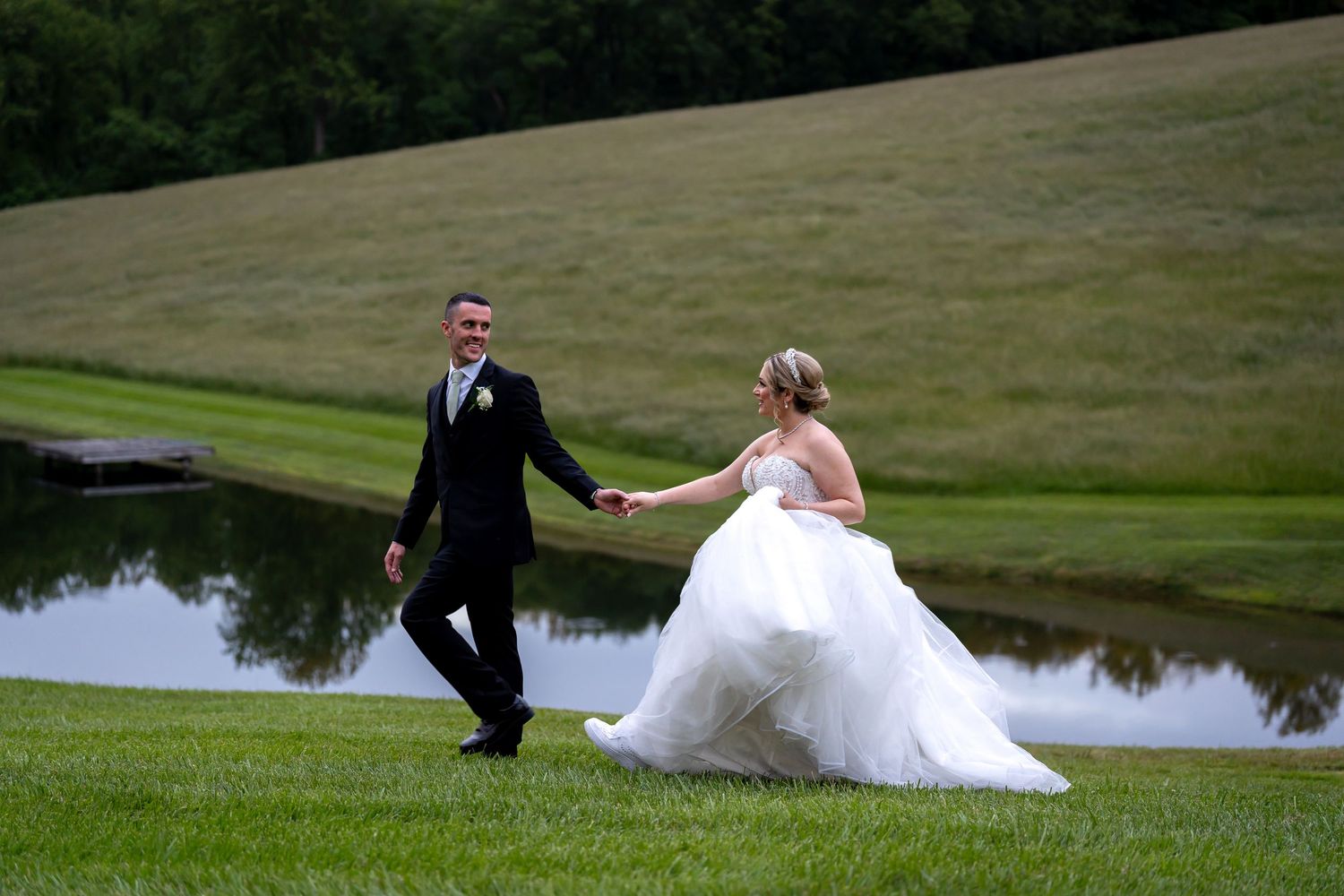 Couple walking hand in hand across a manicured lawn with a pond in the background.