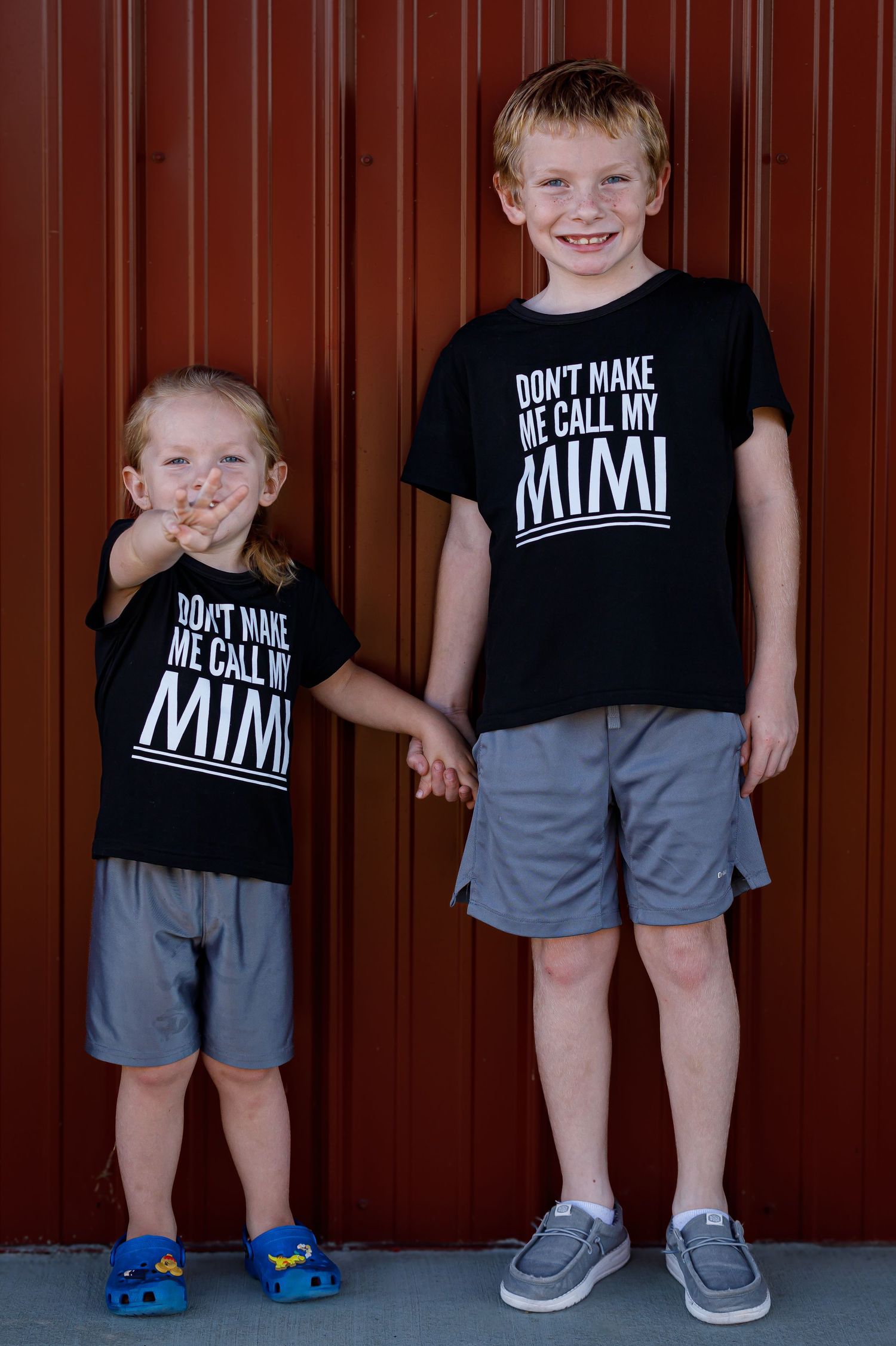Three children stand against red metal wall wearing matching black t-shirts and gray shorts.