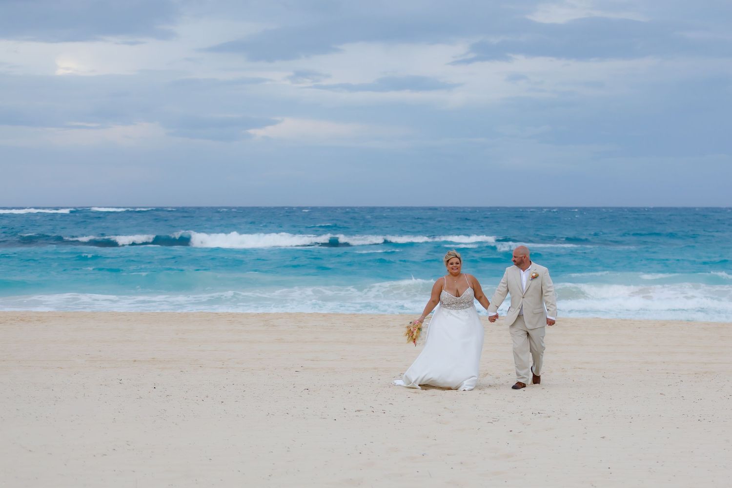 Bride and groom couple walking hand in hand on a tropical beach in Dominican Republic with turquoise waters and cloudy skies.