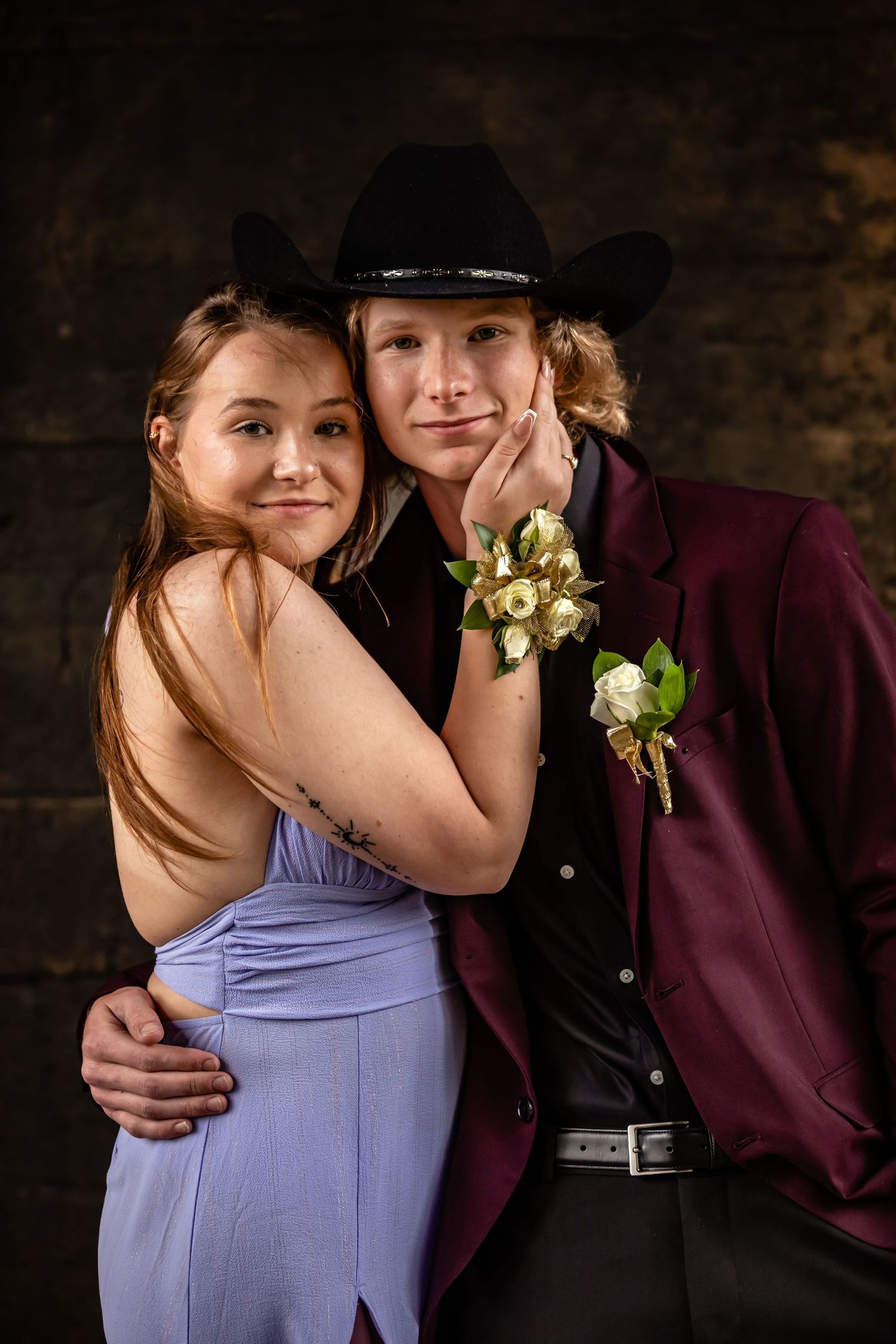 A couple embracing in formal attire with a corsage against a dark backdrop.