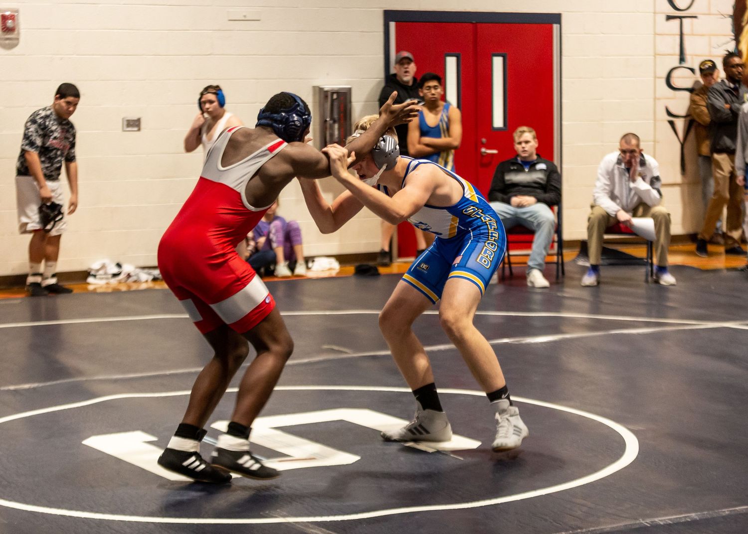 Wrestling match on indoor mat with athletes competing while spectators watch from sidelines.